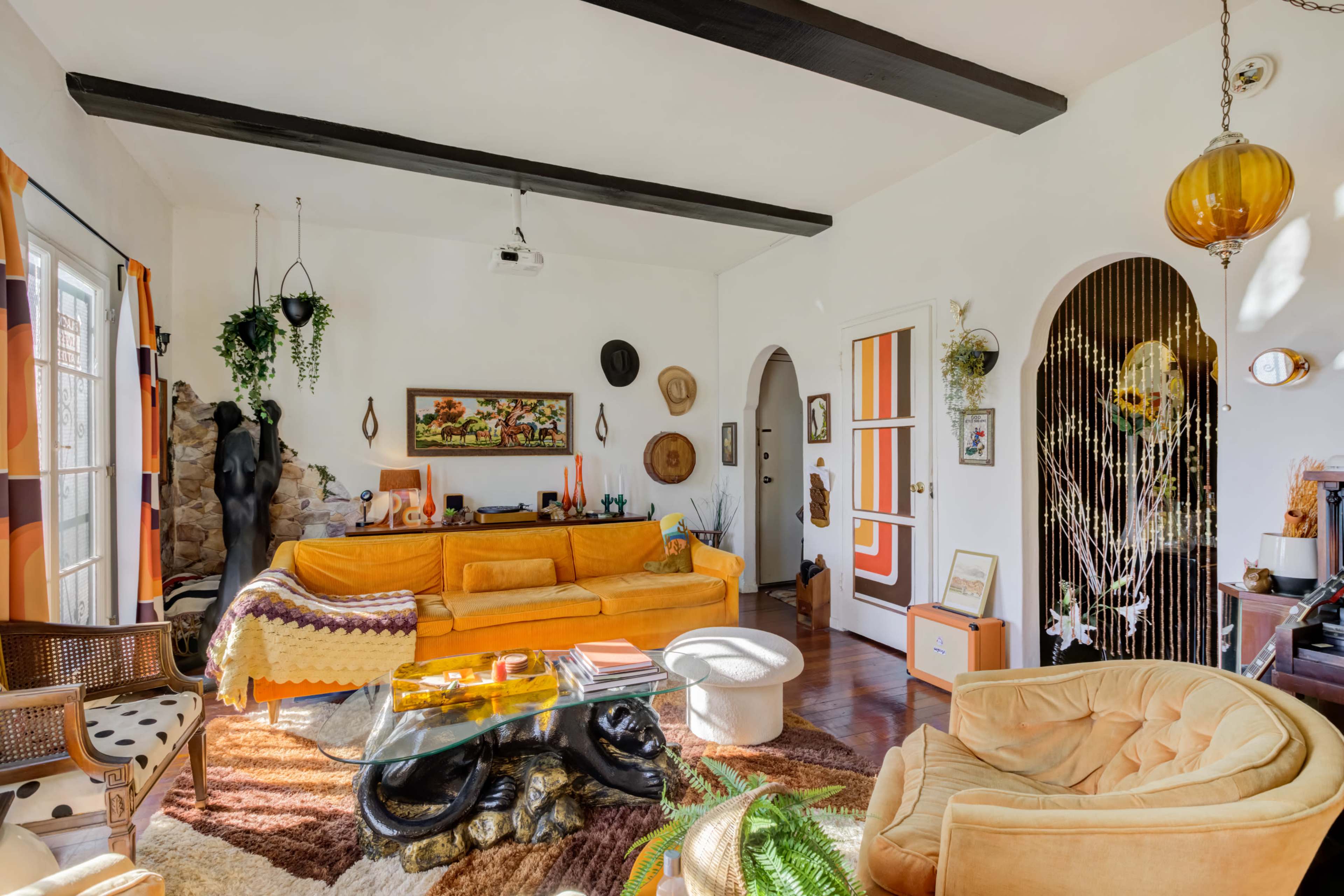 A brightly lit living room featuring orange furniture, a glass coffee table, and decorative plants.