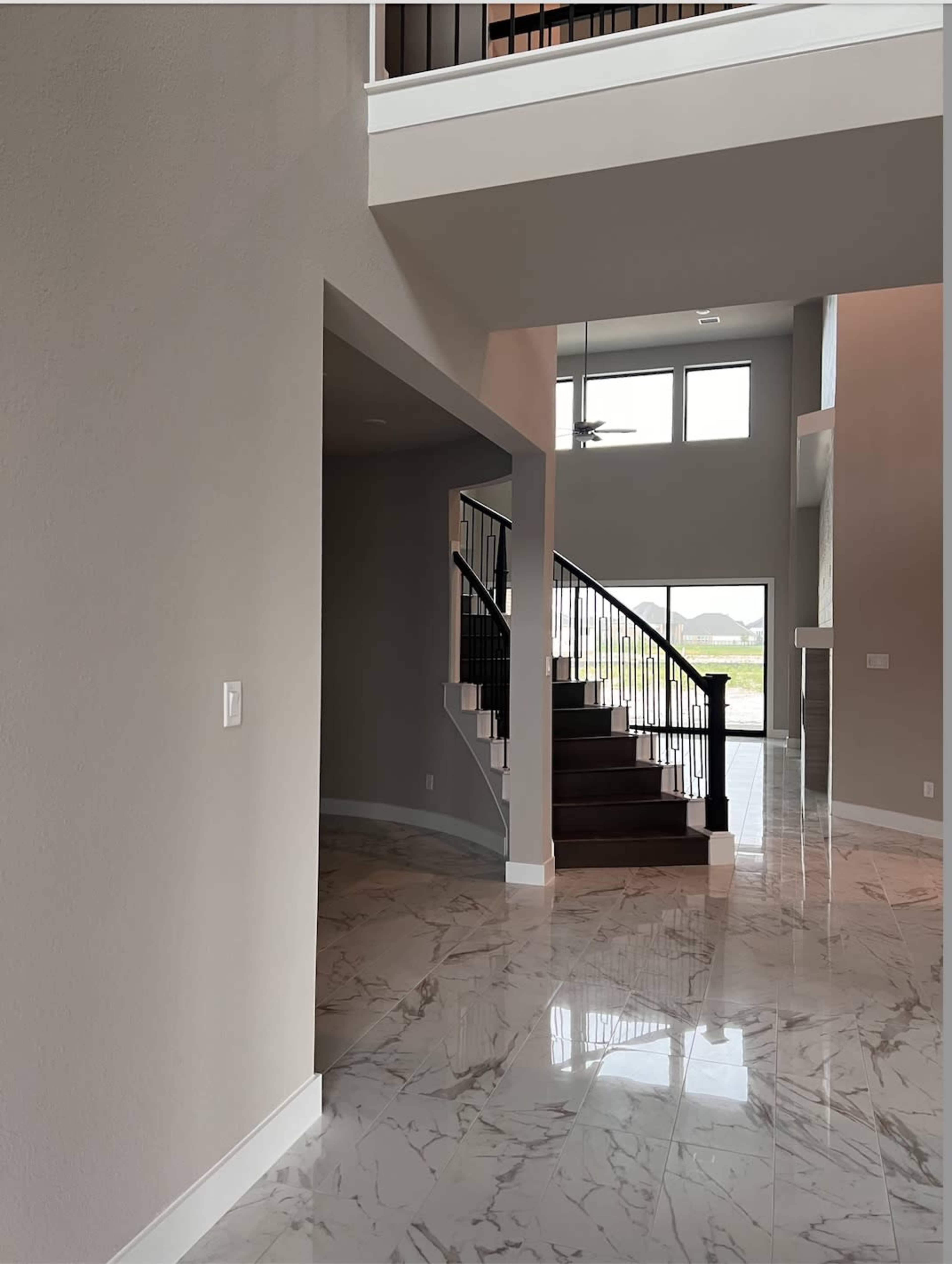 The image depicts the interior of a modern home featuring a spacious entryway with a staircase, polished marble flooring, and large windows providing natural light.