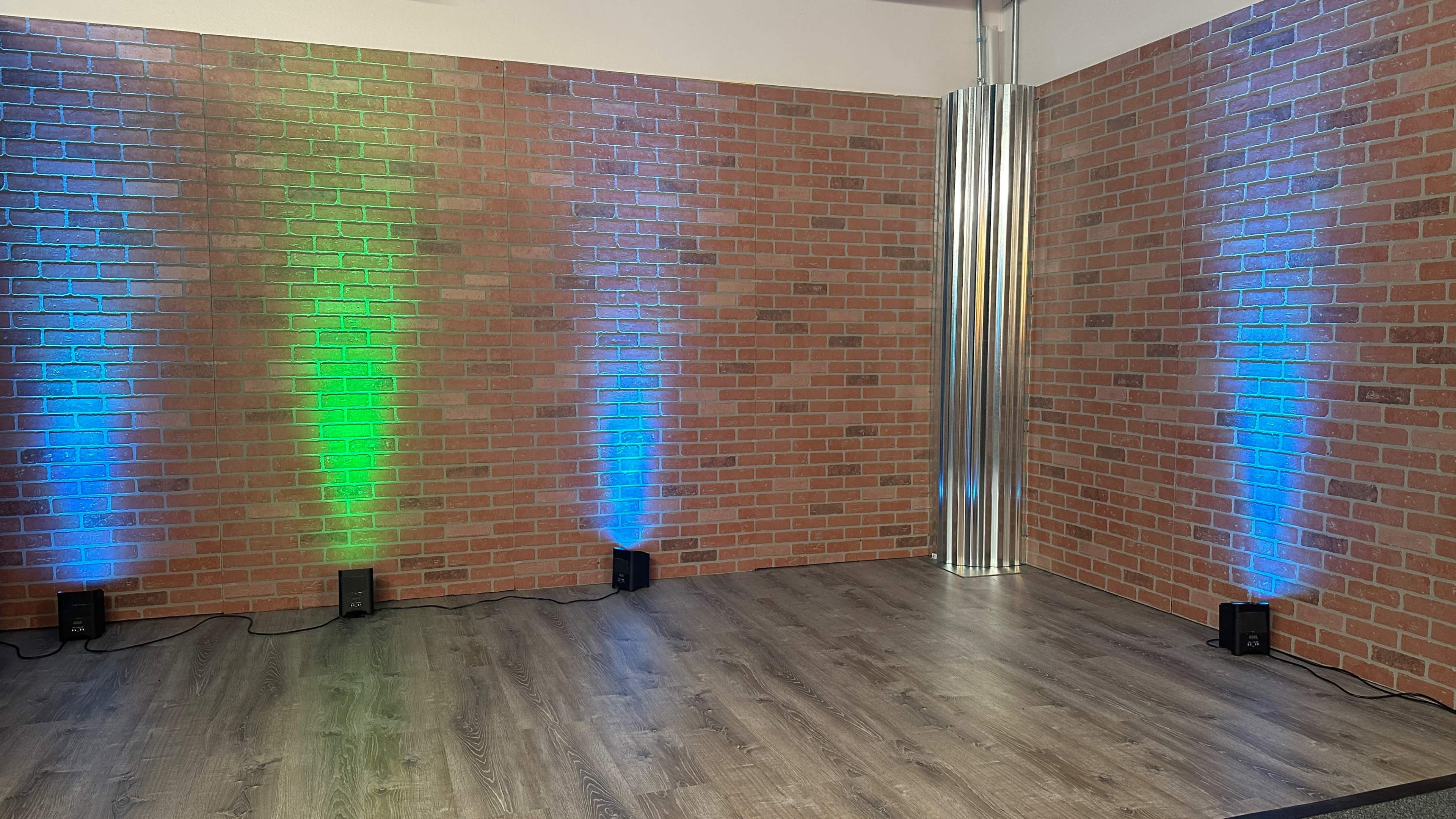 The image shows a corner of a room with a brick wall, lit by blue and green lights, and featuring a wooden floor.