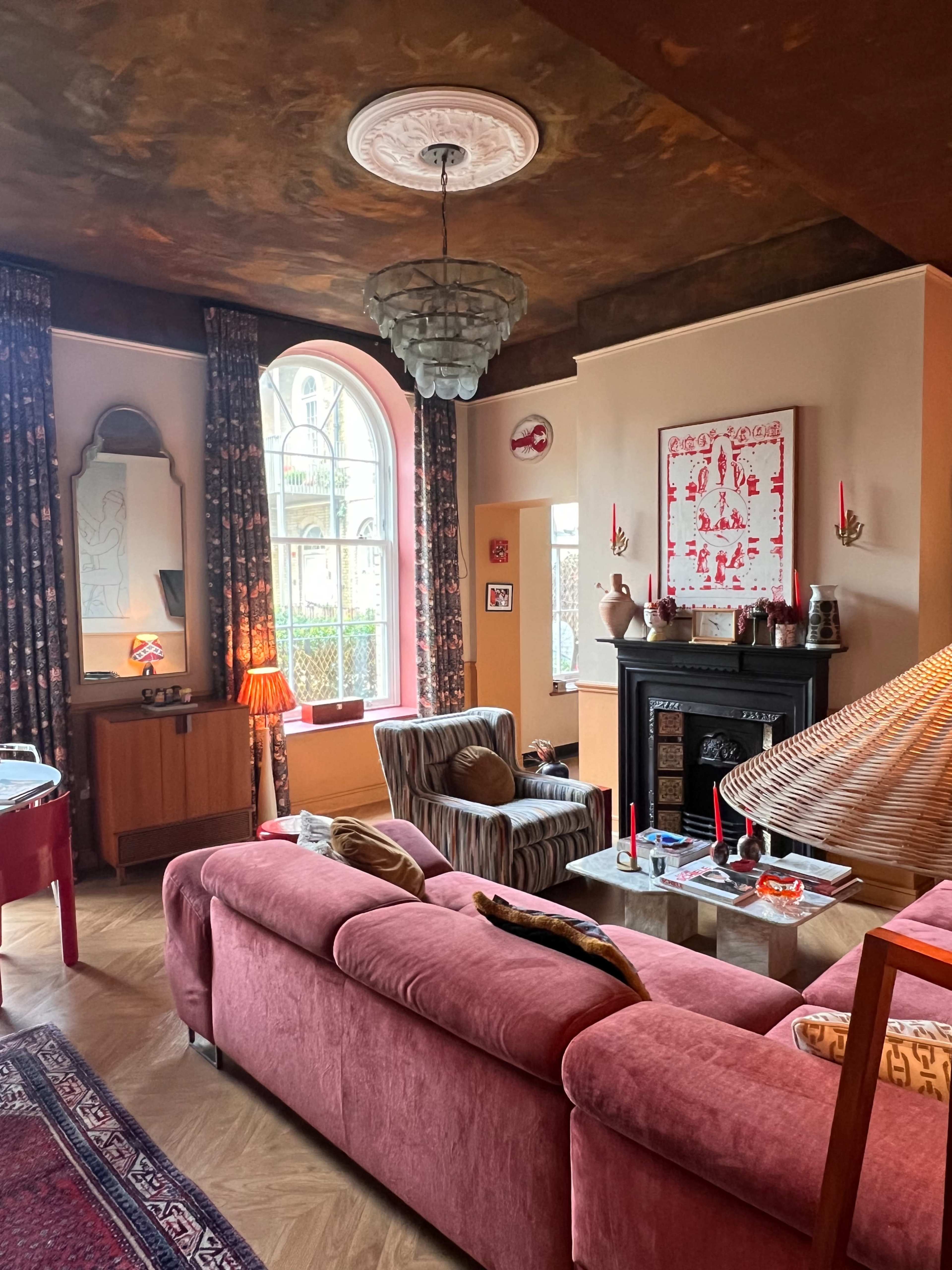 A cozy living room features a pink sofa, a patterned armchair, and a fireplace, with large windows allowing natural light to illuminate the space.
