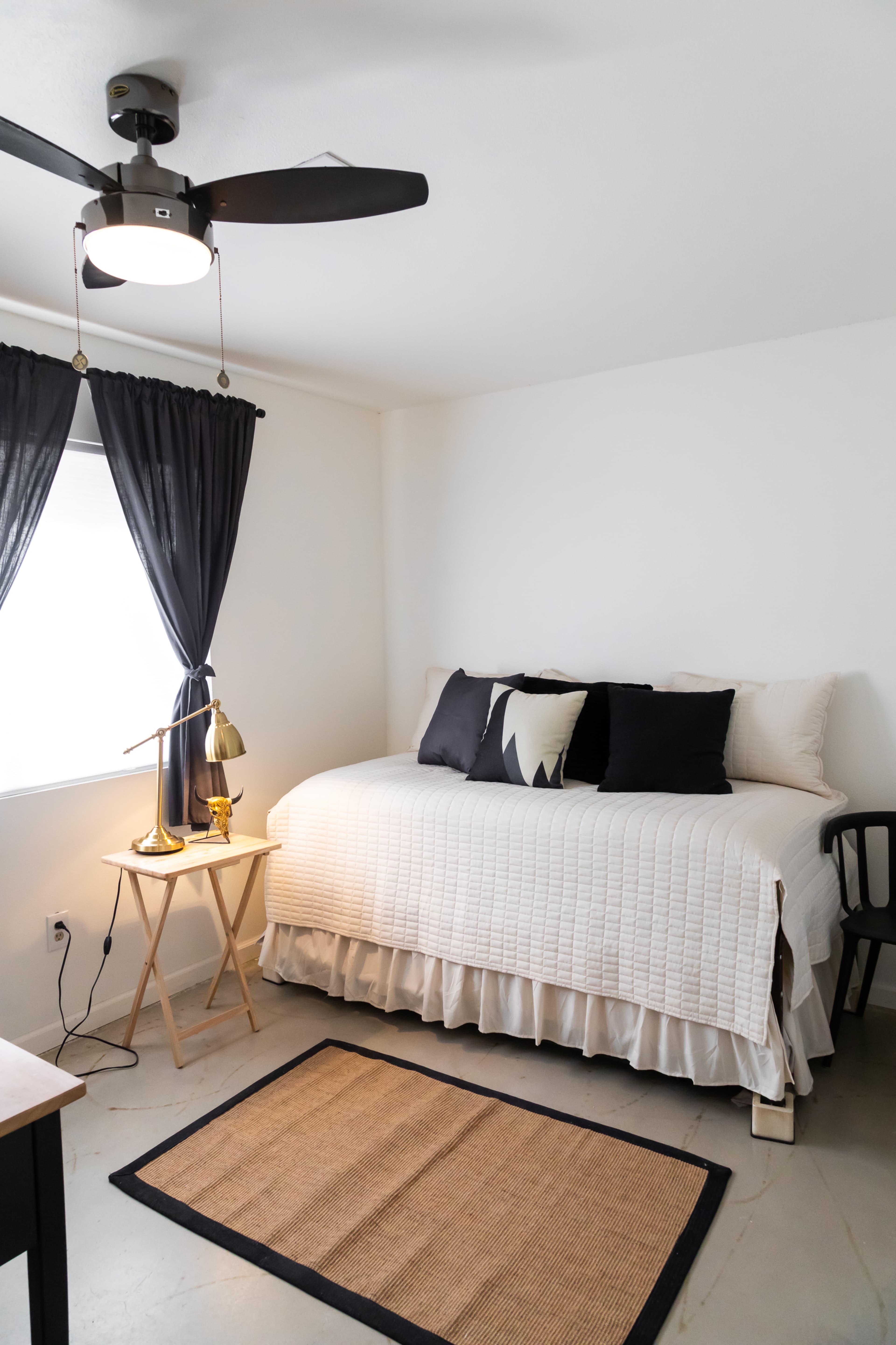 A neatly arranged bedroom featuring a daybed with a white quilt, black pillows, a small bedside table with a lamp, and a woven rug on the floor.