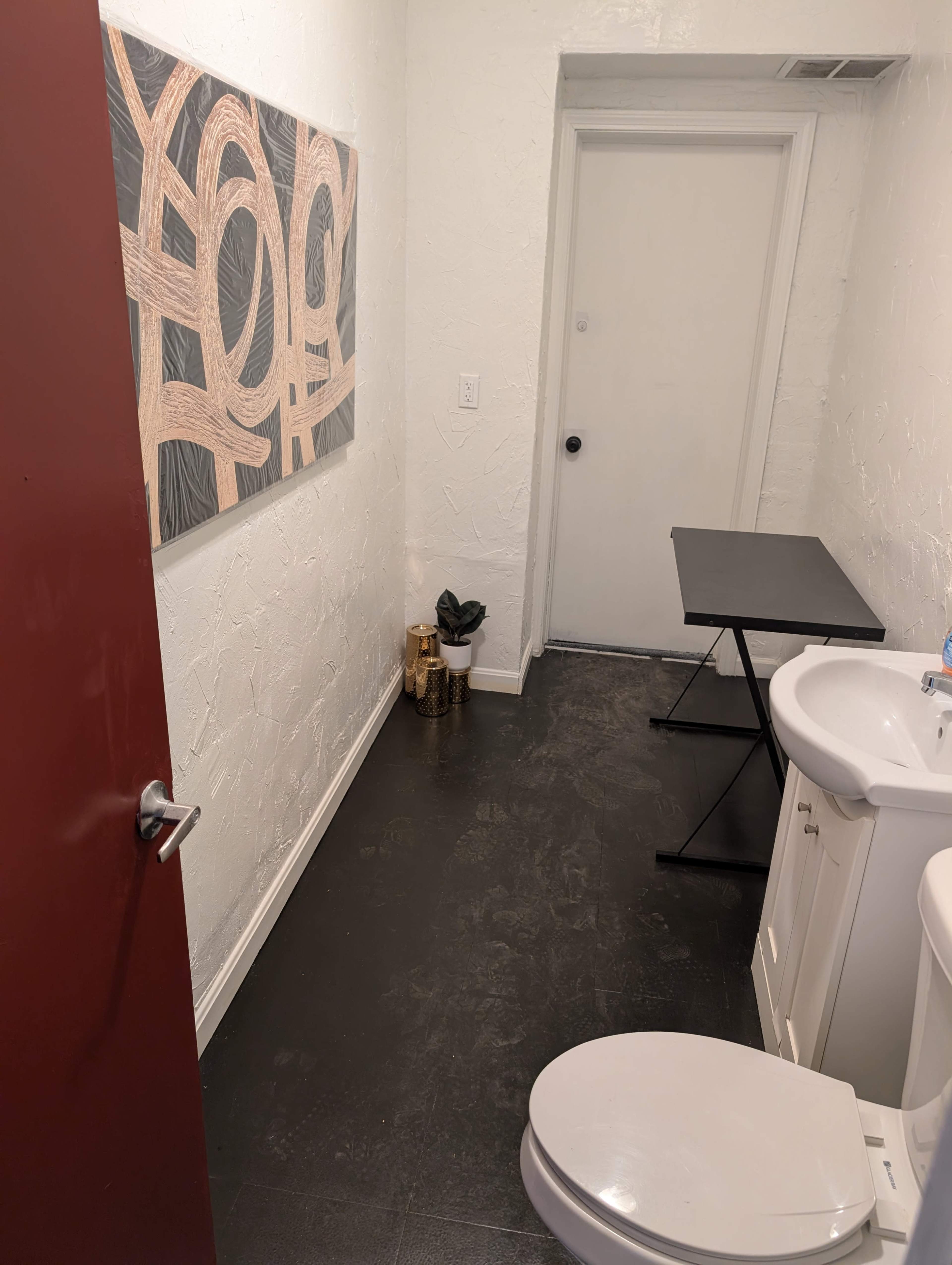 The image shows a small bathroom with two sinks, a toilet, a black table, and a door leading to another room, along with abstract artwork on the wall.