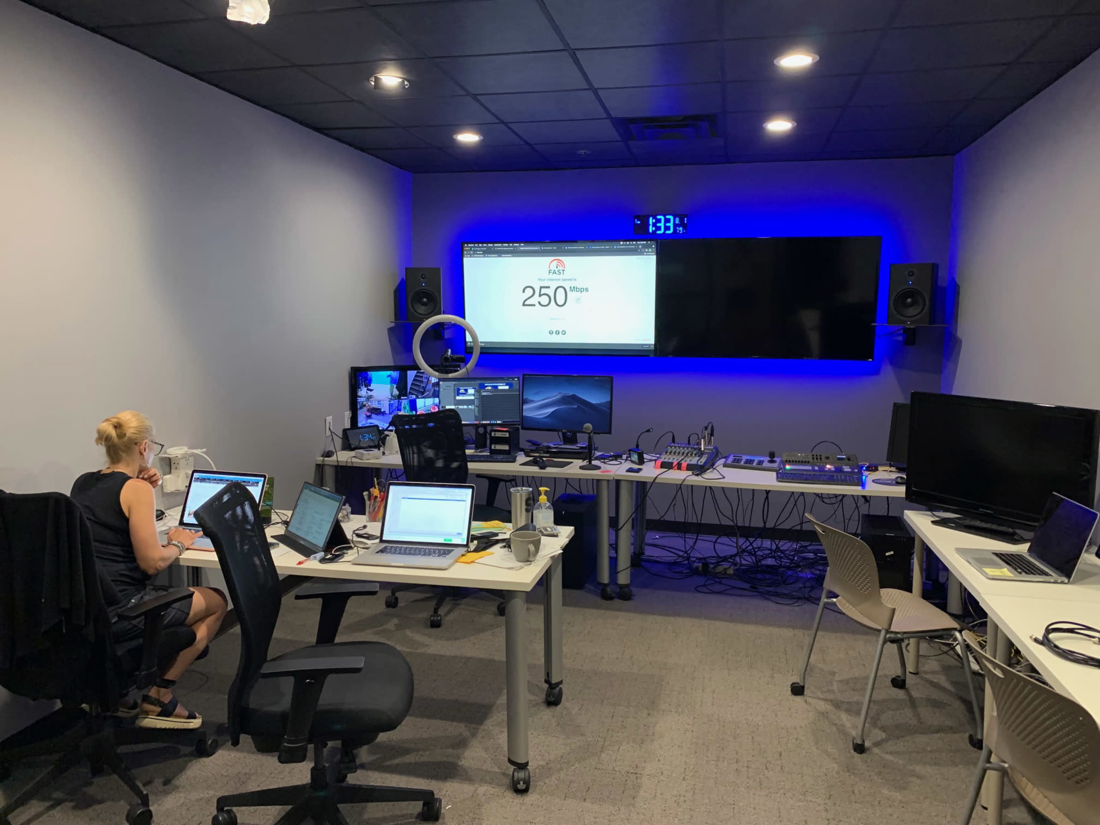 A modern office space features multiple workstations with computers, a large screen displaying a website, and blue ambient lighting.