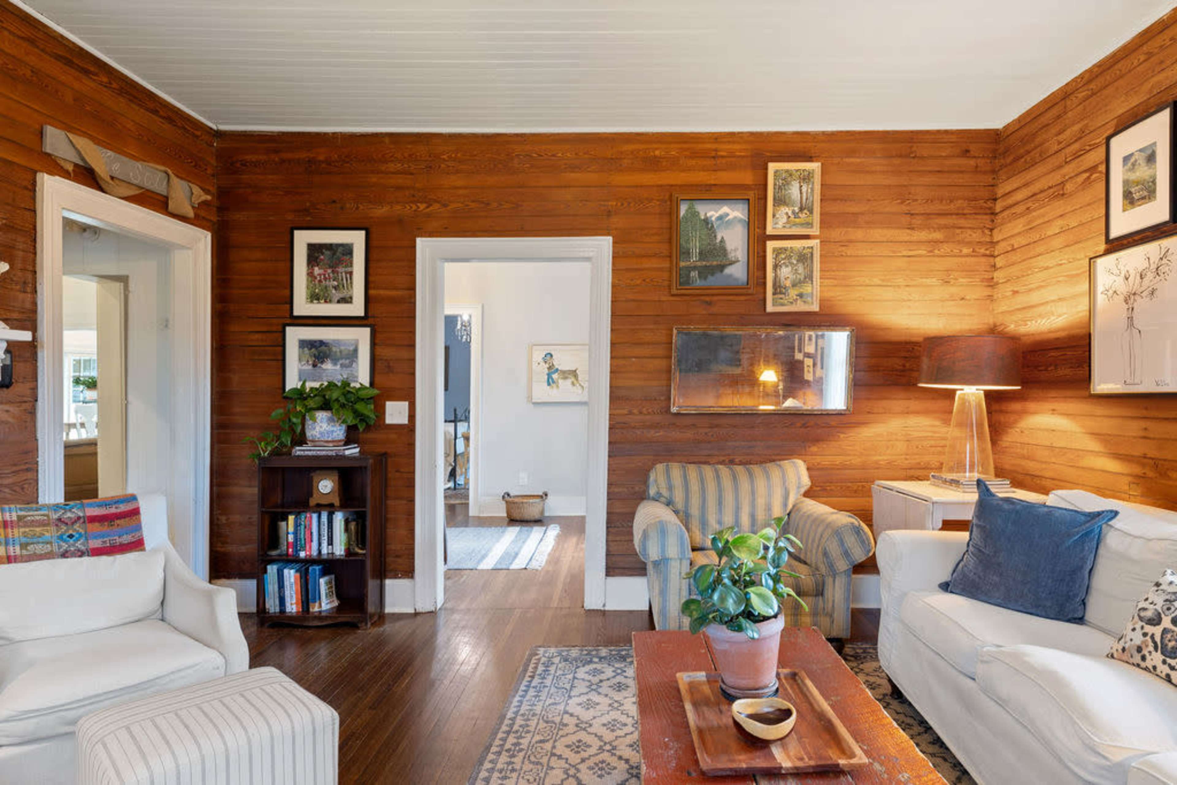 The living room features wood-paneled walls, two sofas, a wooden coffee table, and a bookshelf, with a doorway leading to another room in the background.