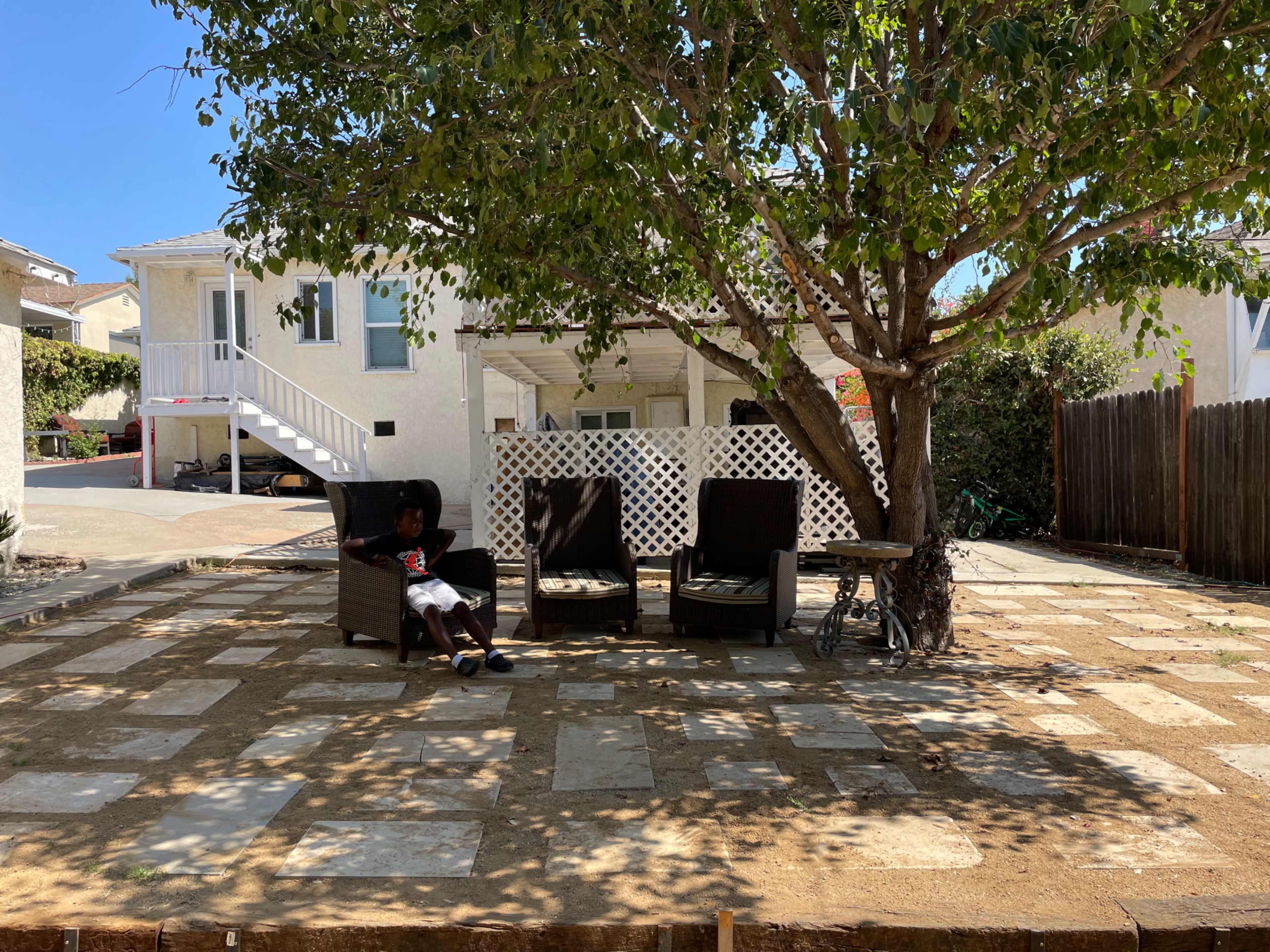 A person sits on a chair under a tree in a patio area with stone paving and a residential house in the background.