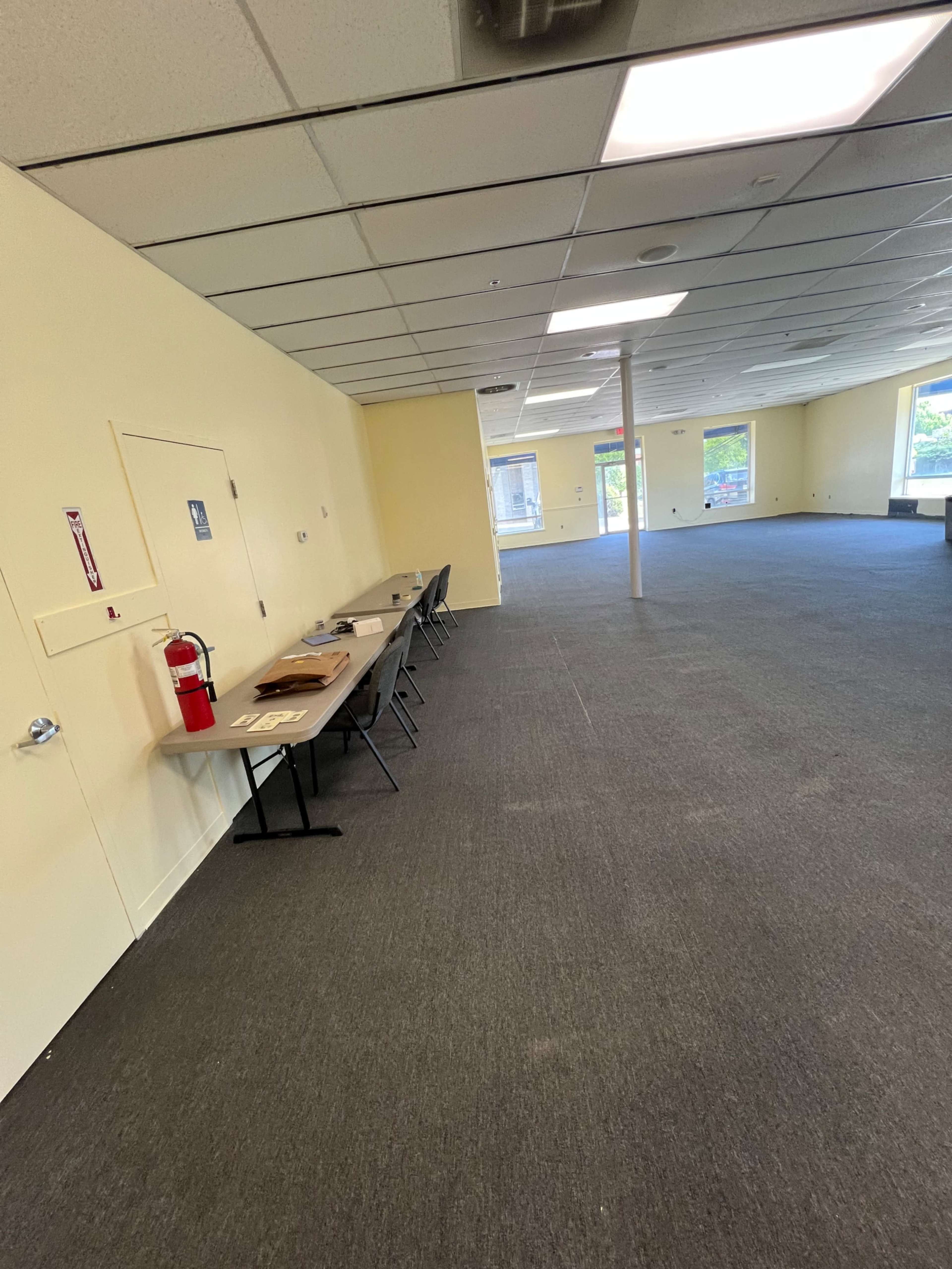 The image shows an empty room with beige walls and a gray carpet, featuring a row of tables along one wall and a door on the left side.