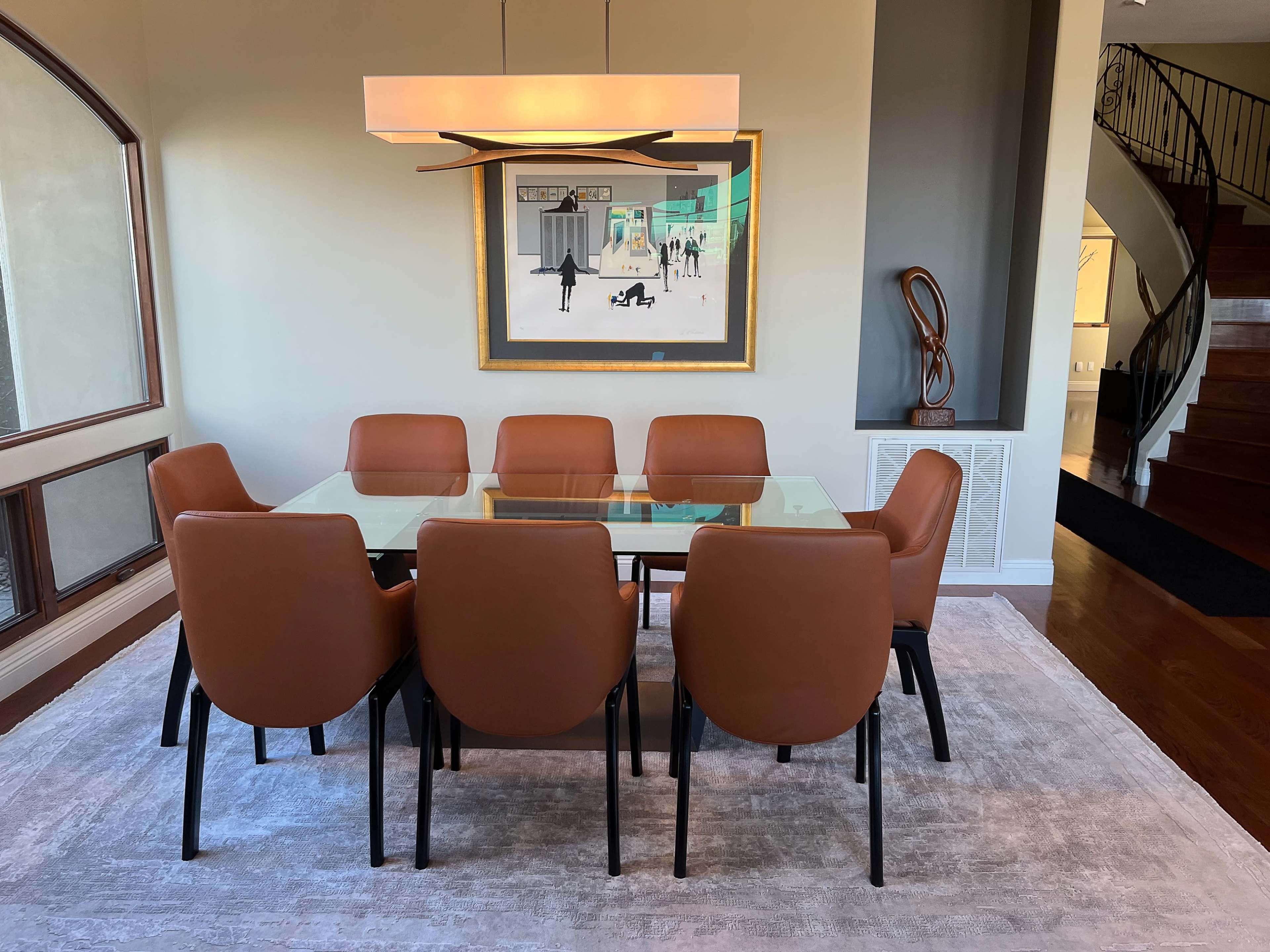 A spacious dining room features a glass table surrounded by eight brown leather chairs, with an artistic painting on the wall and a modern light fixture above.