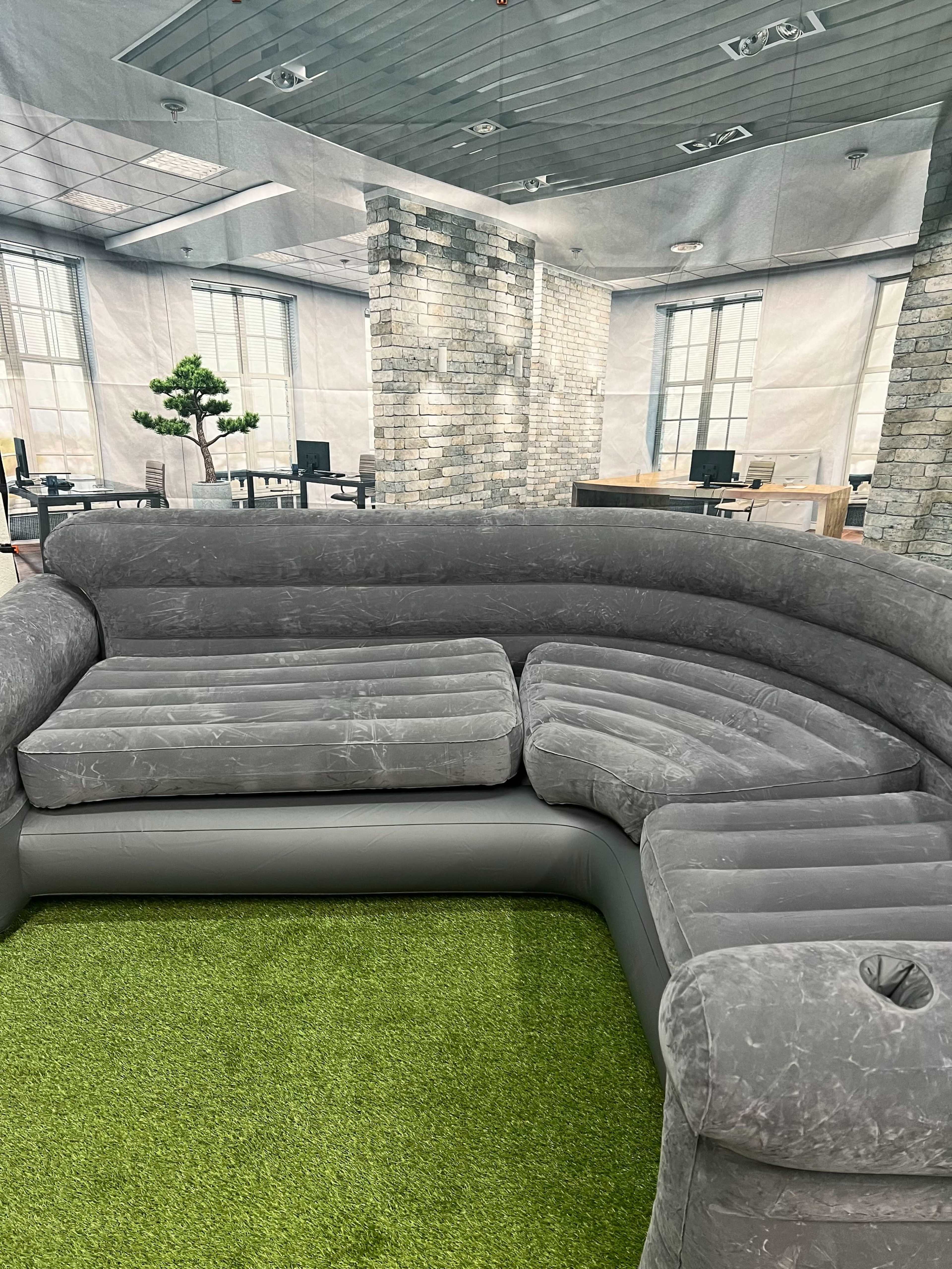 A gray inflatable couch situated on artificial grass in a modern office space with brick walls and visible workstations in the background.