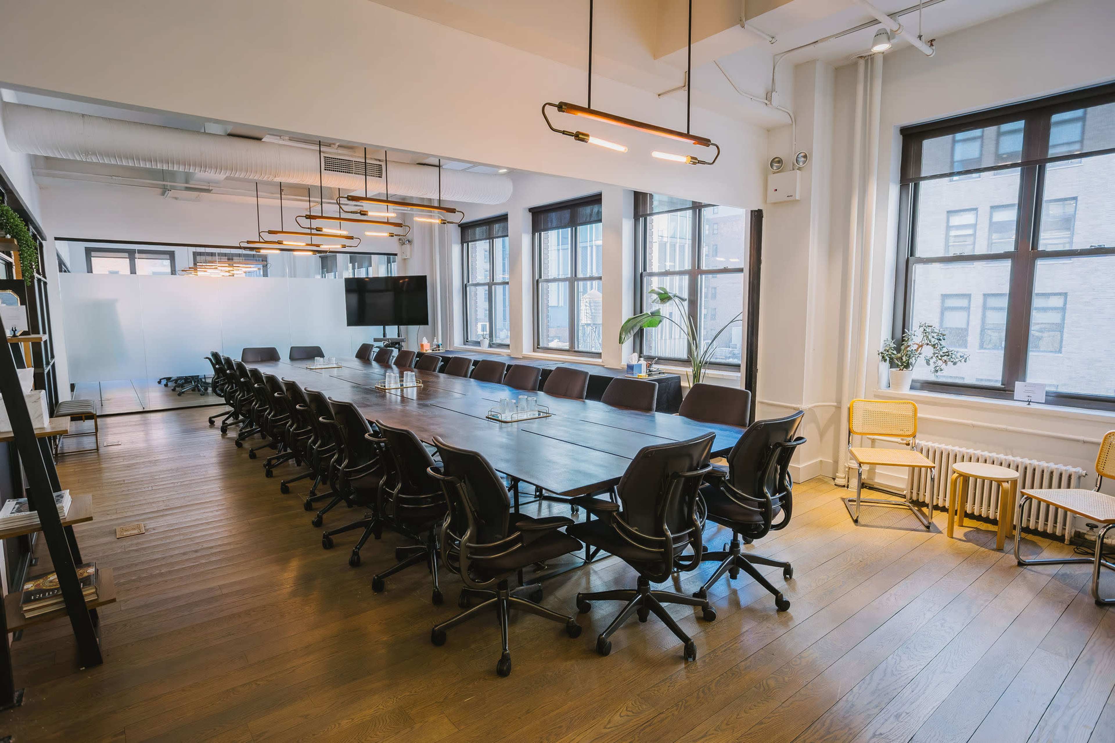 A modern conference room features a long table surrounded by black office chairs, with large windows allowing natural light to fill the space.