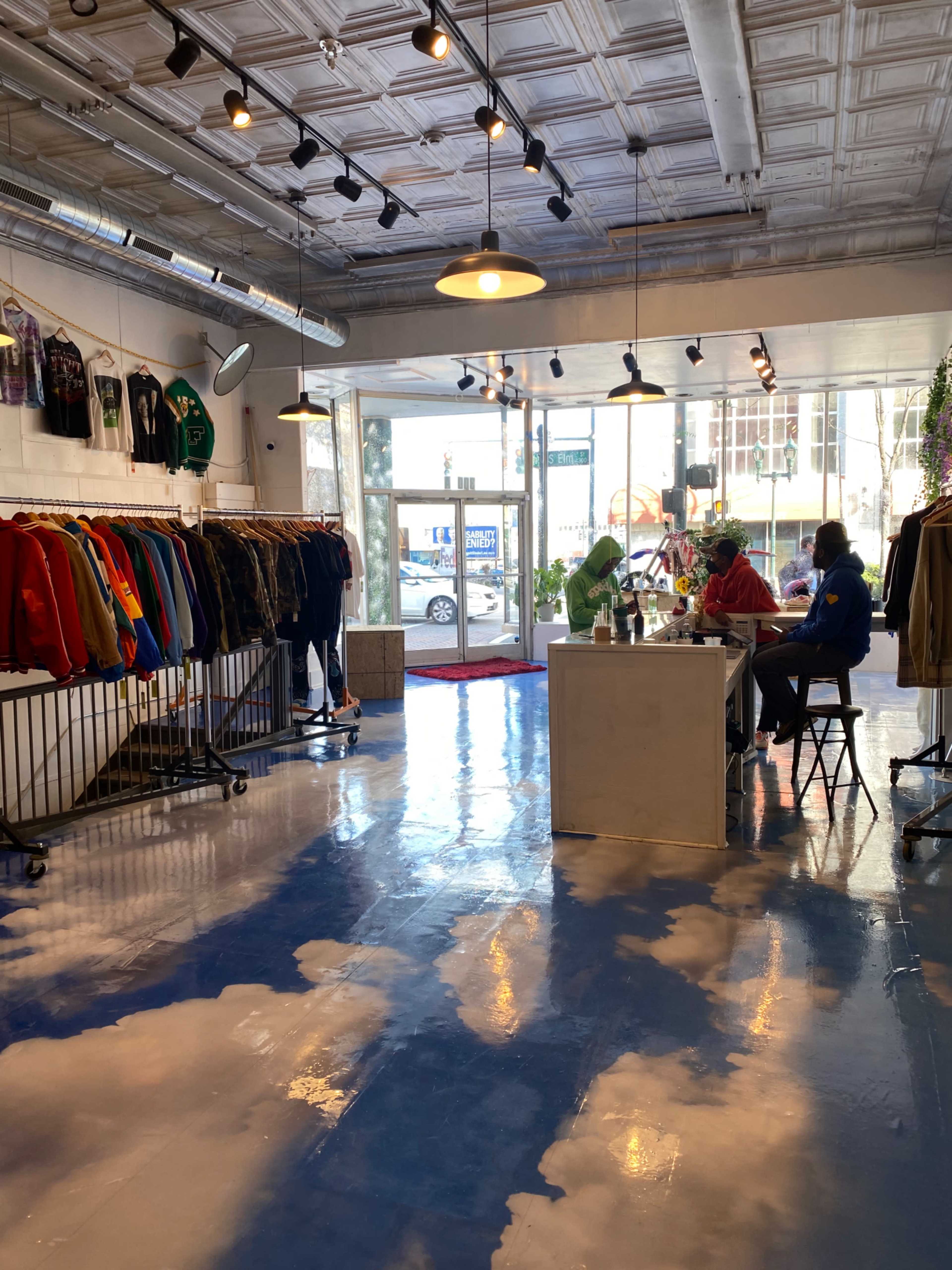 A clothing store with racks of colorful apparel on one side and two people seated at a counter on the opposite side, against a blue floor resembling a sky with clouds.
