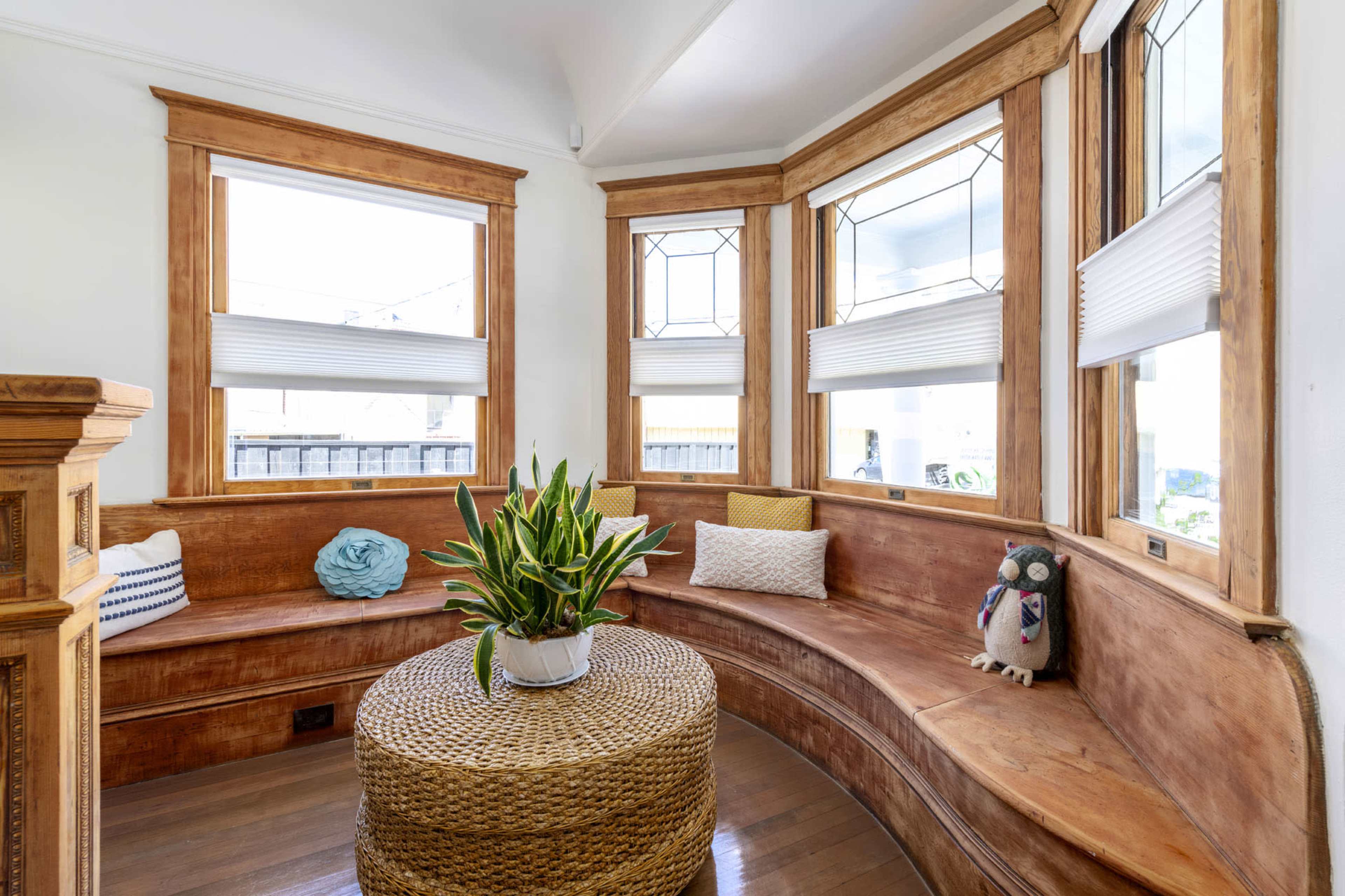 A cozy alcove features a wooden bench, a round woven coffee table, and potted plants, with large windows allowing natural light to enter.
