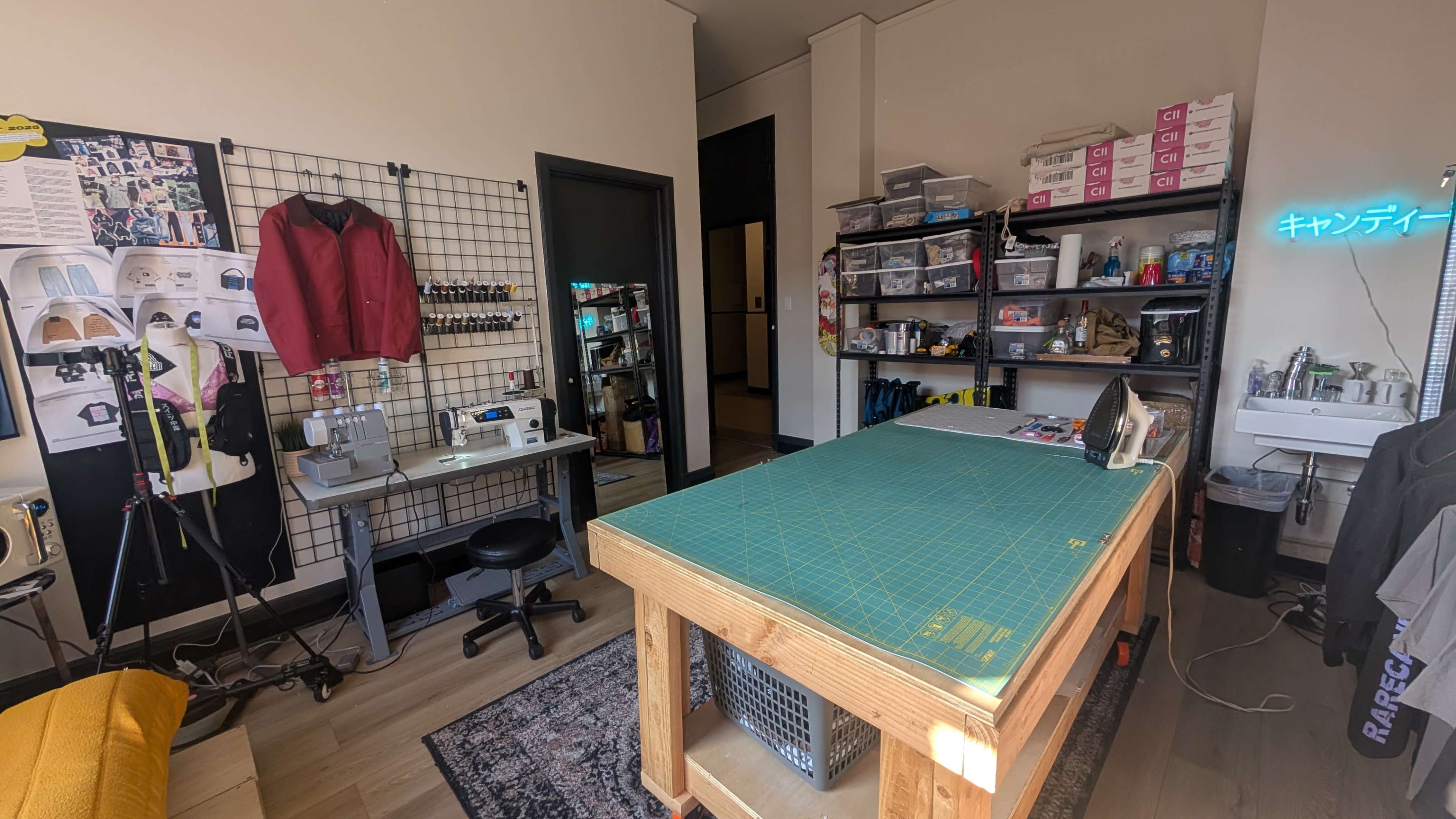 The image shows a sewing workspace featuring a large cutting table, a sewing machine, and organized shelving with various supplies.