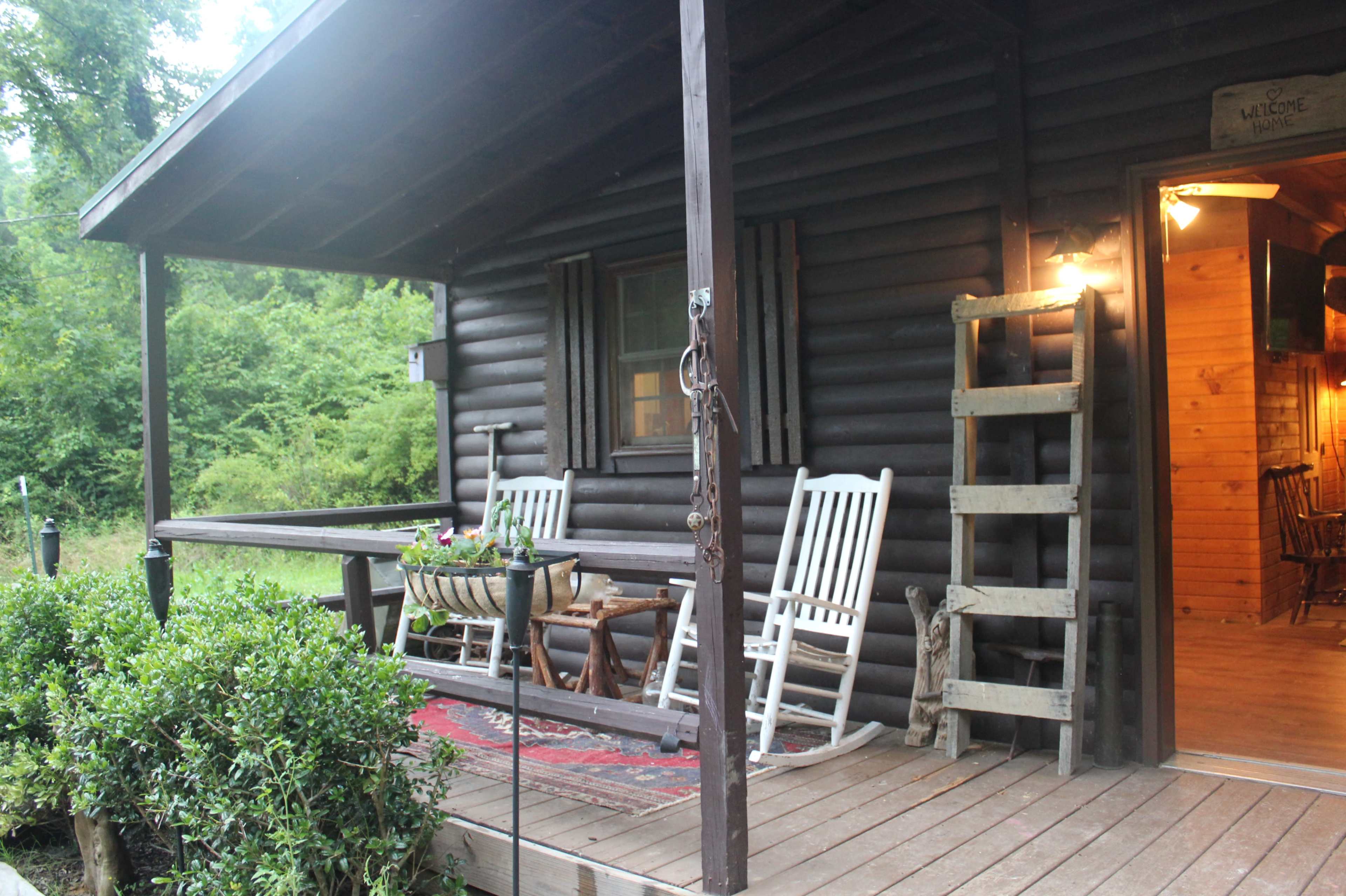 A log cabin features a porch with rocking chairs, a rug, and a wooden ladder, surrounded by greenery.