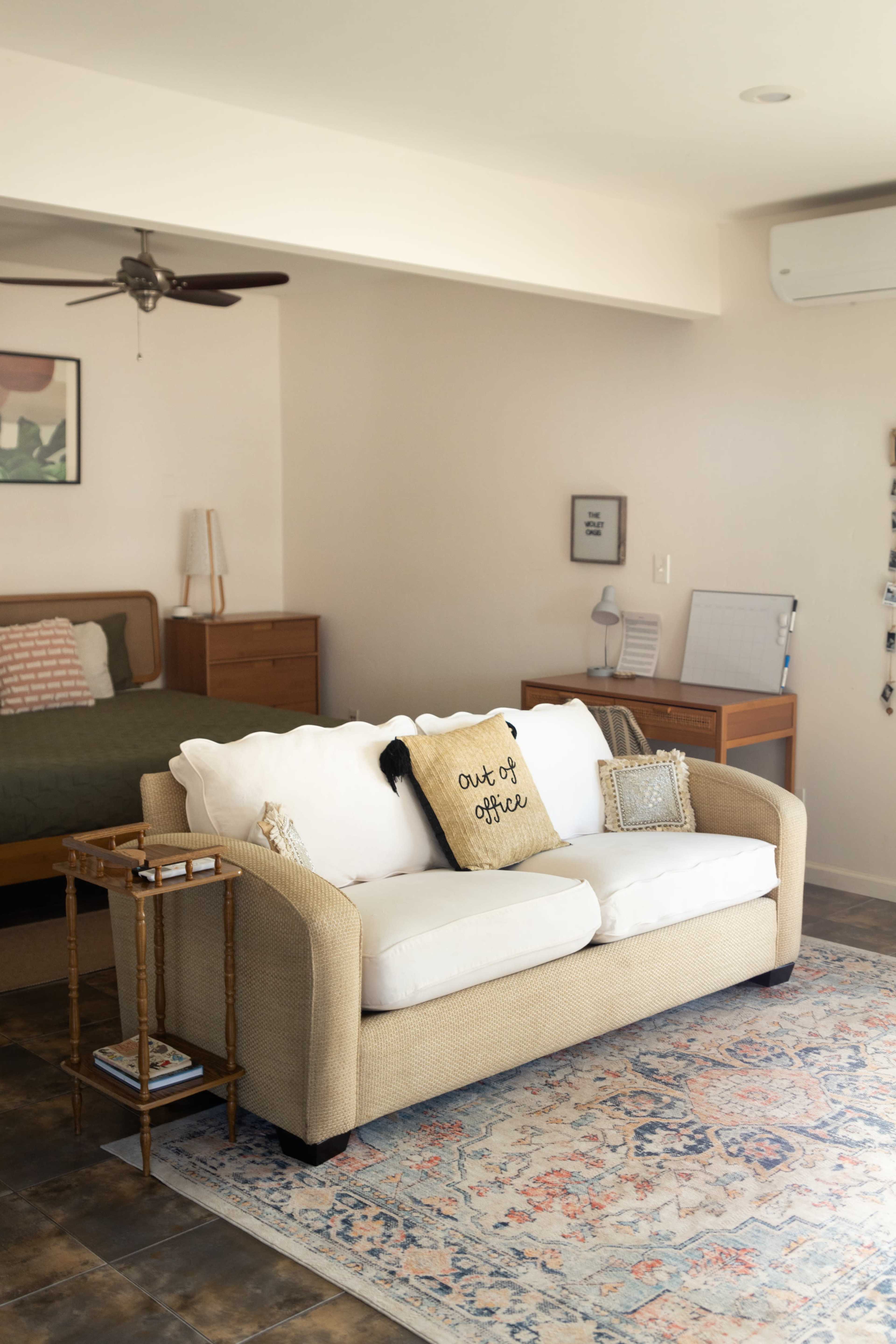 A cozy living space features a beige couch with white cushions, a small side table, a bed in the background, and wooden furniture accents.