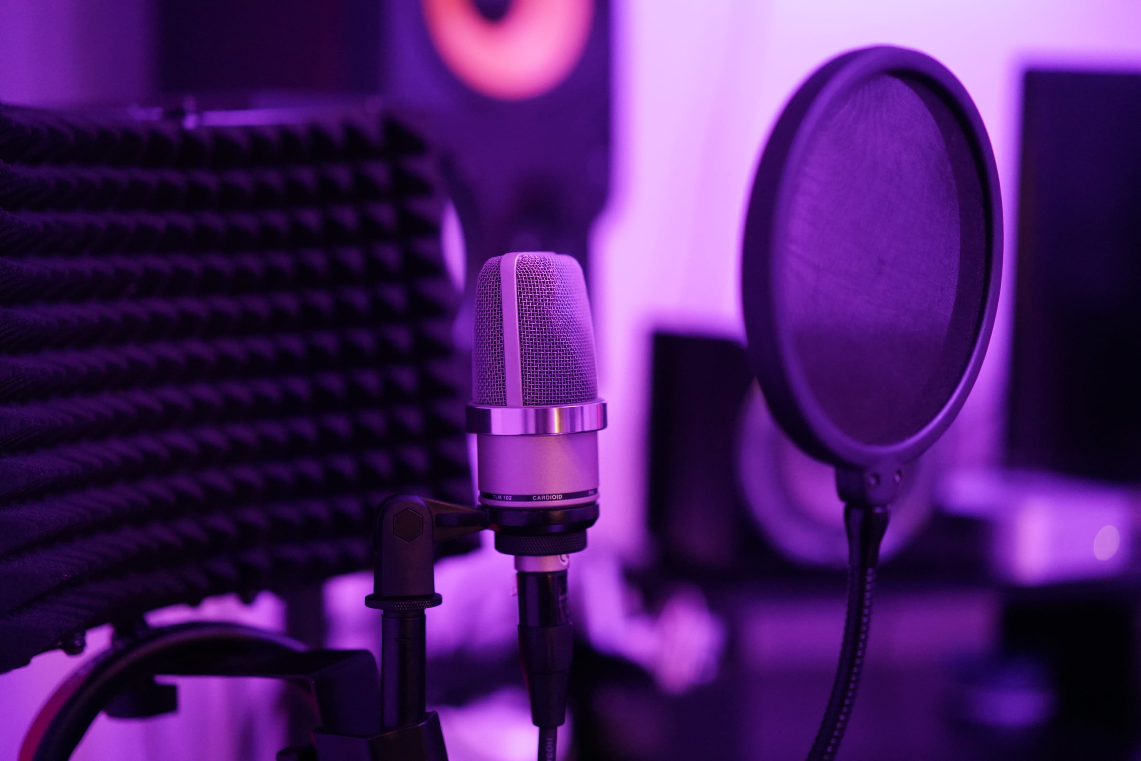 The image features a microphone positioned near a pop filter, with soundproofing panels and speakers in the background under purple lighting.