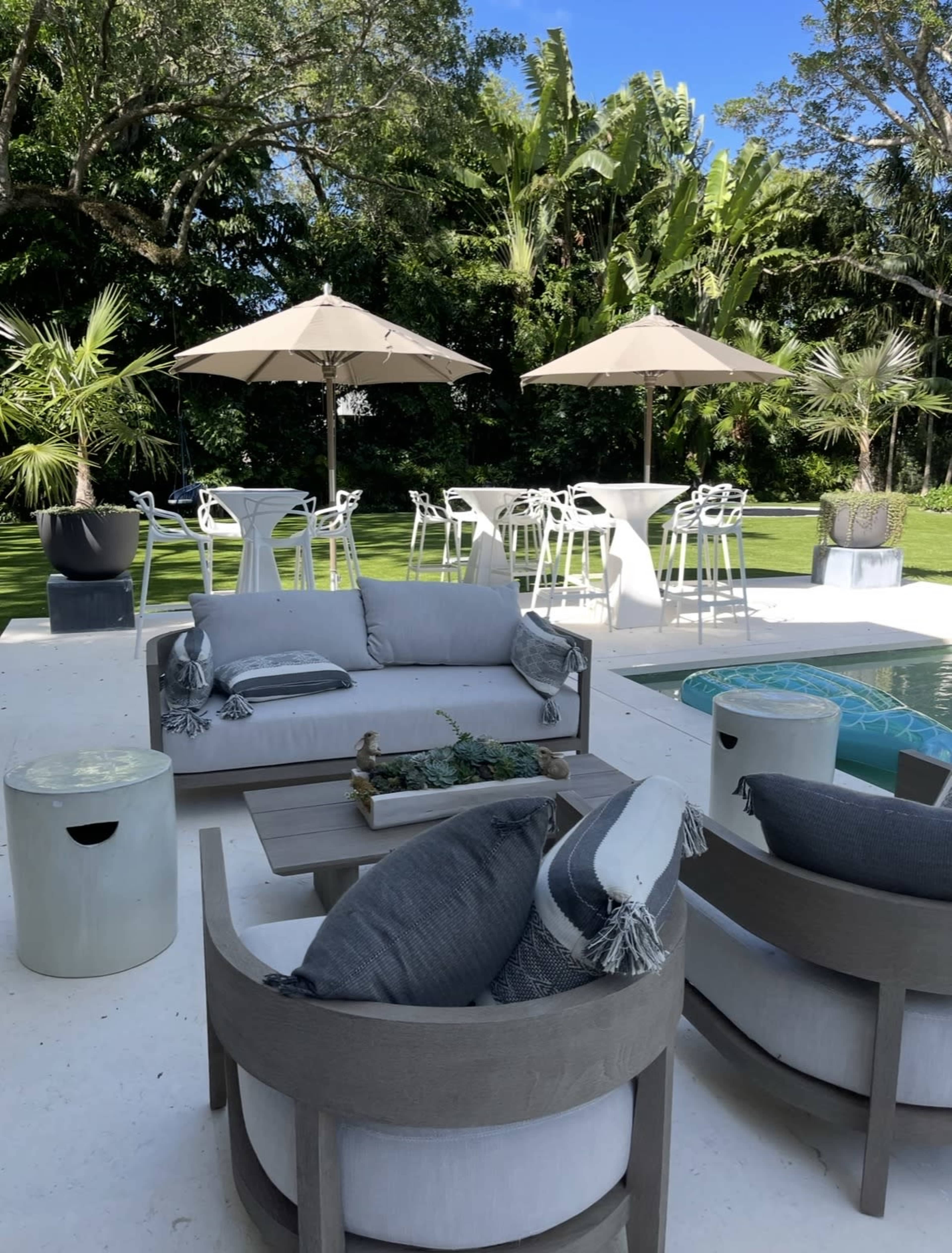 The image shows an outdoor patio area with modern furniture, two large umbrellas, a swimming pool, and lush greenery in the background.