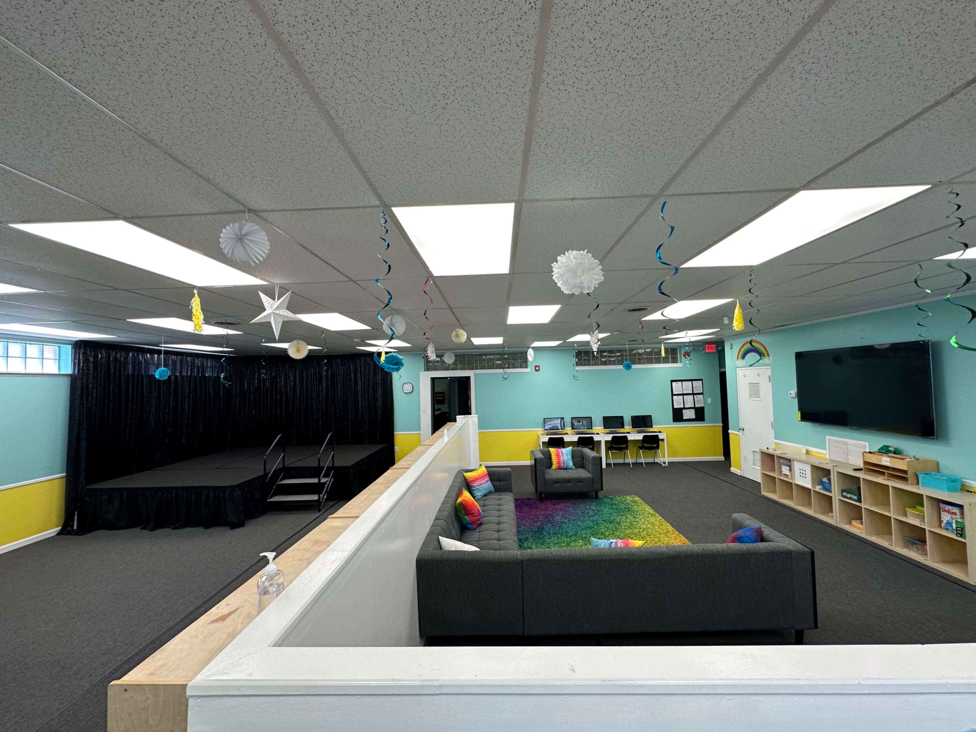 A bright, spacious room with a seating area featuring a gray couch, a small stage at one end, and colorful decorations hanging from the ceiling.