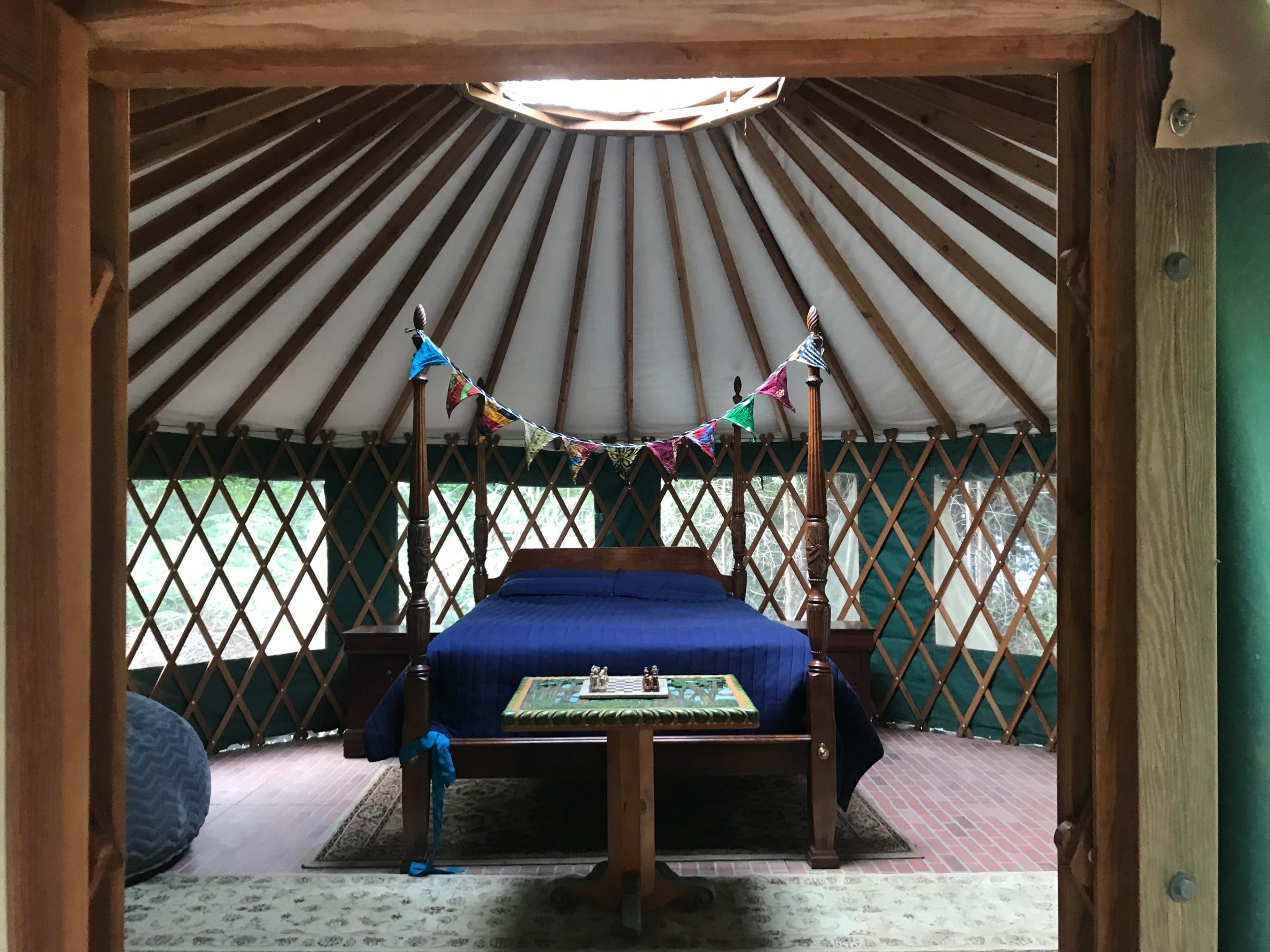 A round yurt interior features a bed at the center, with colorful decorations hanging above and windows that provide a view of the outdoors.