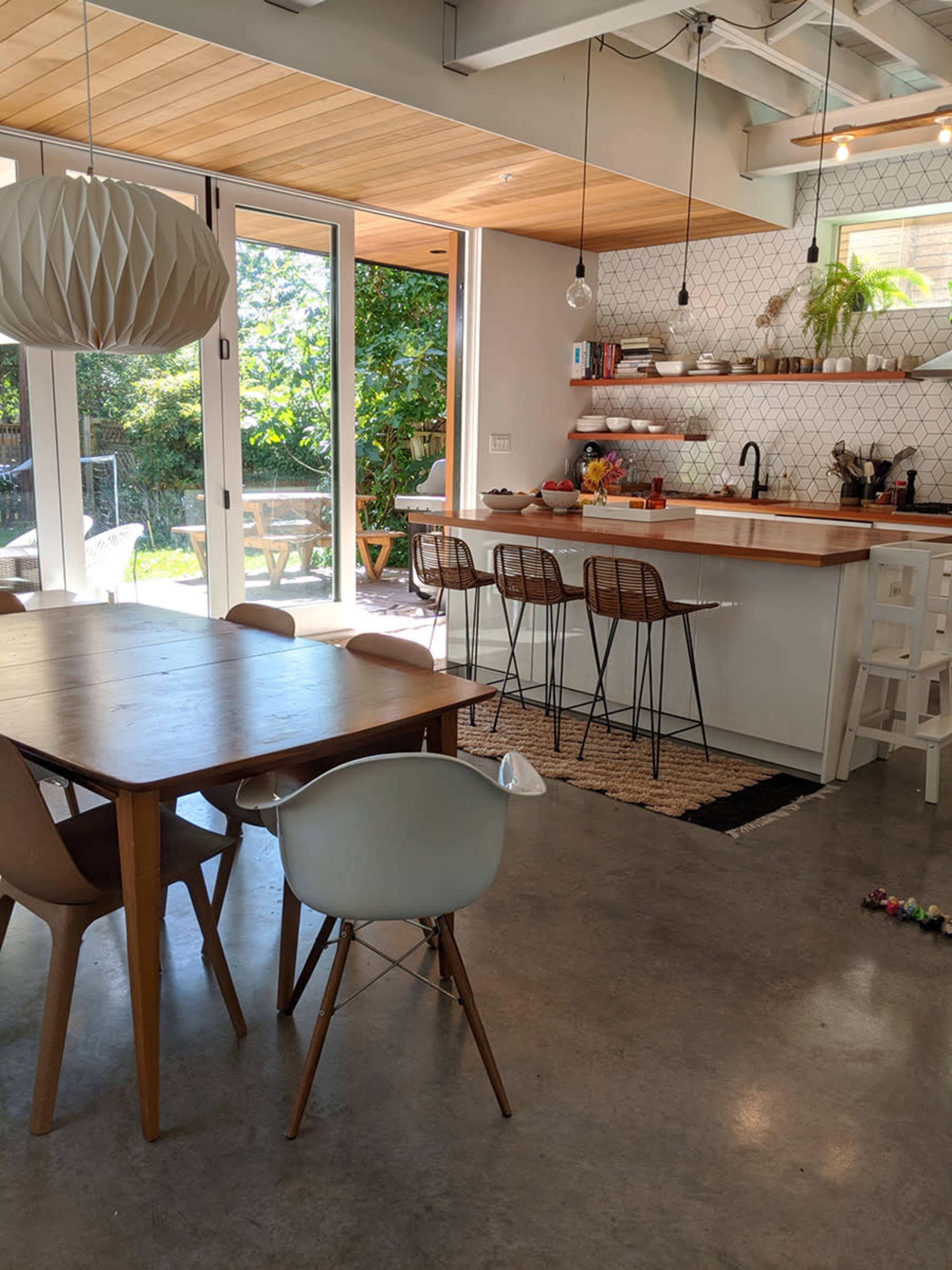 A modern kitchen and dining area features a wooden table, bentwood chairs, a long counter with stools, and large windows offering a view of the garden.
