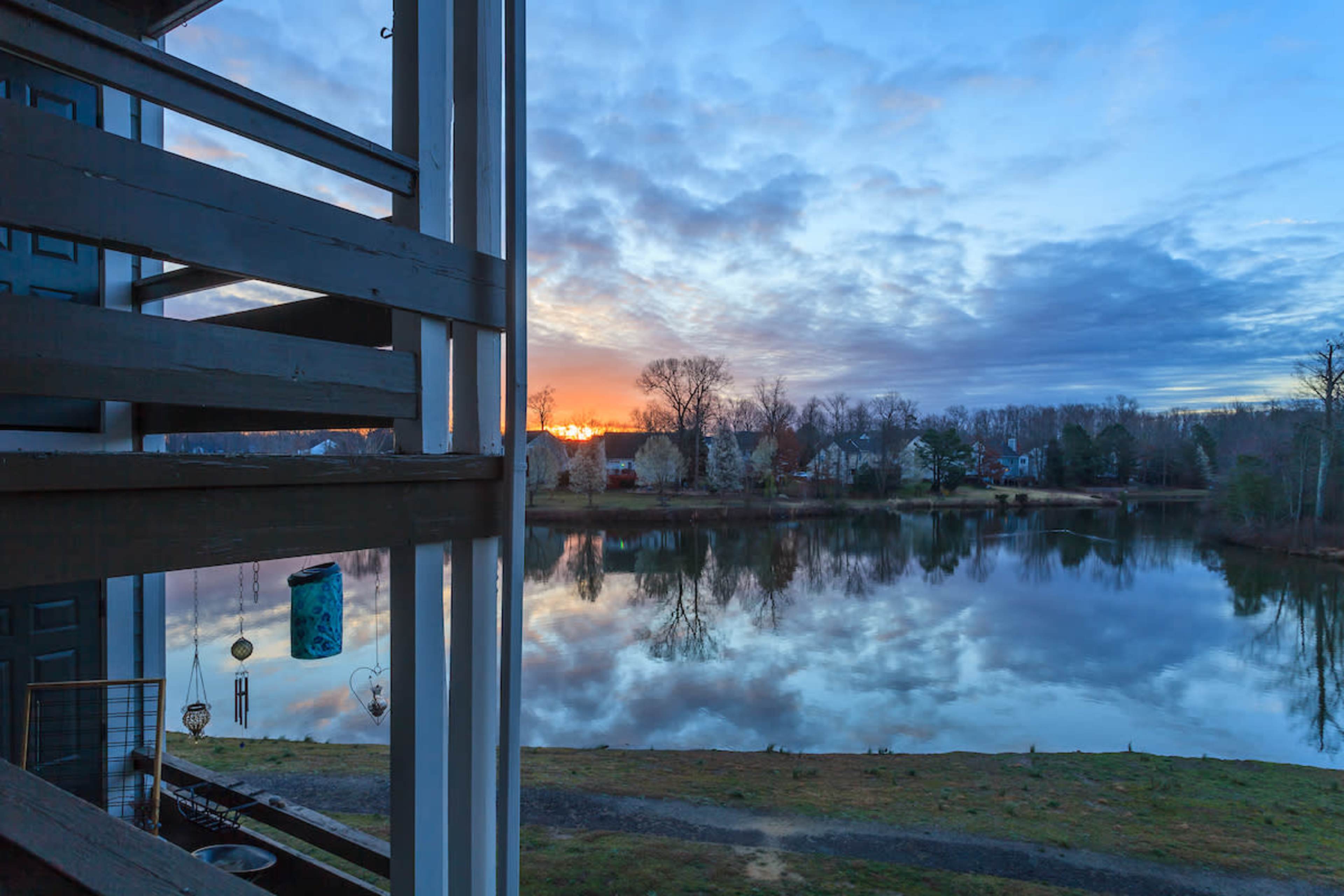 The image shows a calm lake reflecting a colorful sunset, framed by trees and a building balcony in the foreground.