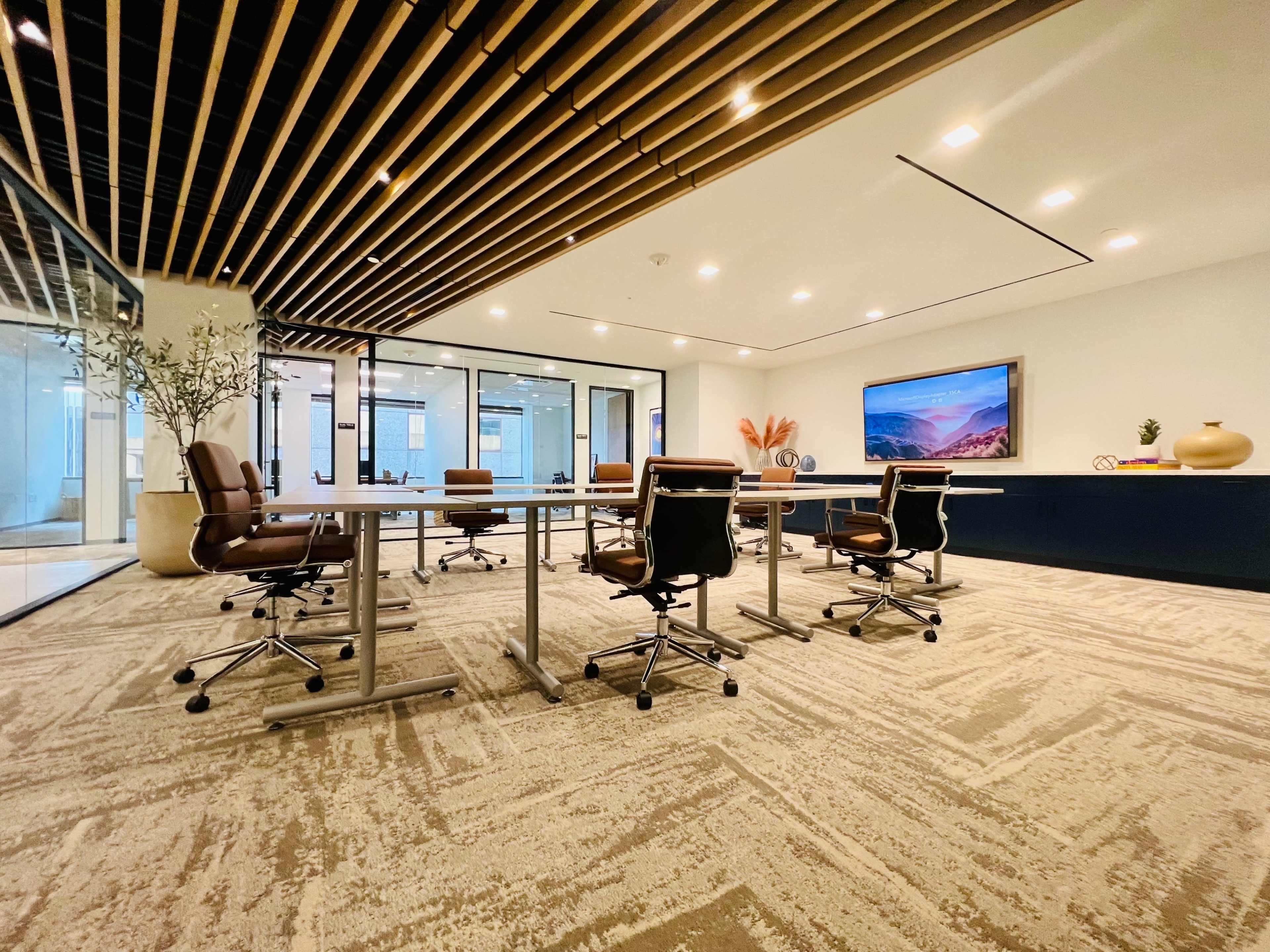 A modern conference room features a large table surrounded by ergonomic chairs, with a wall-mounted screen and glass partitions.