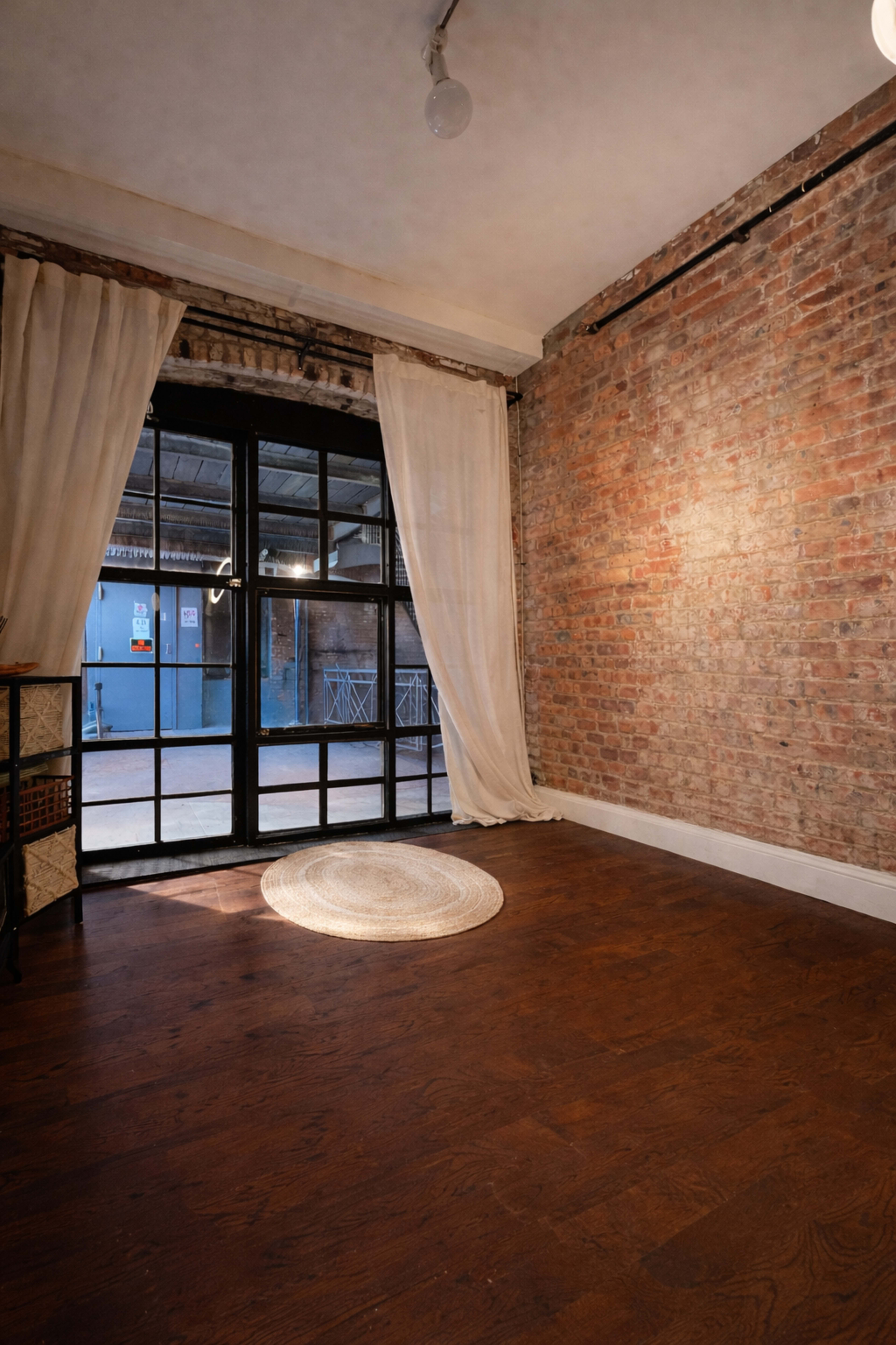 The image shows a room with exposed brick walls, large windows covered with sheer curtains, and hardwood flooring with a round rug in the center.
