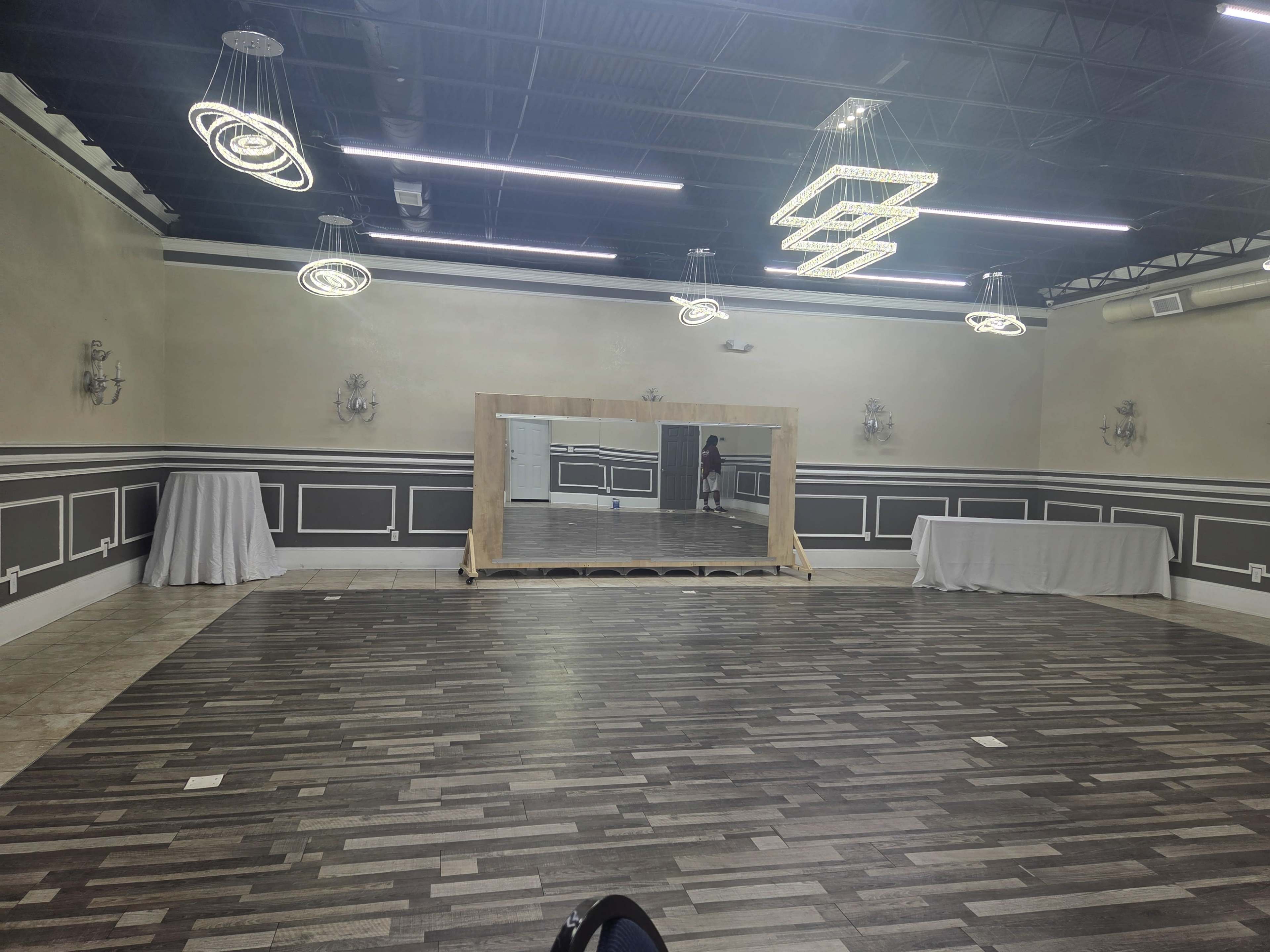 The image shows a spacious room with wooden flooring, a large mirror on a wooden stand, and modern light fixtures hanging from the ceiling.