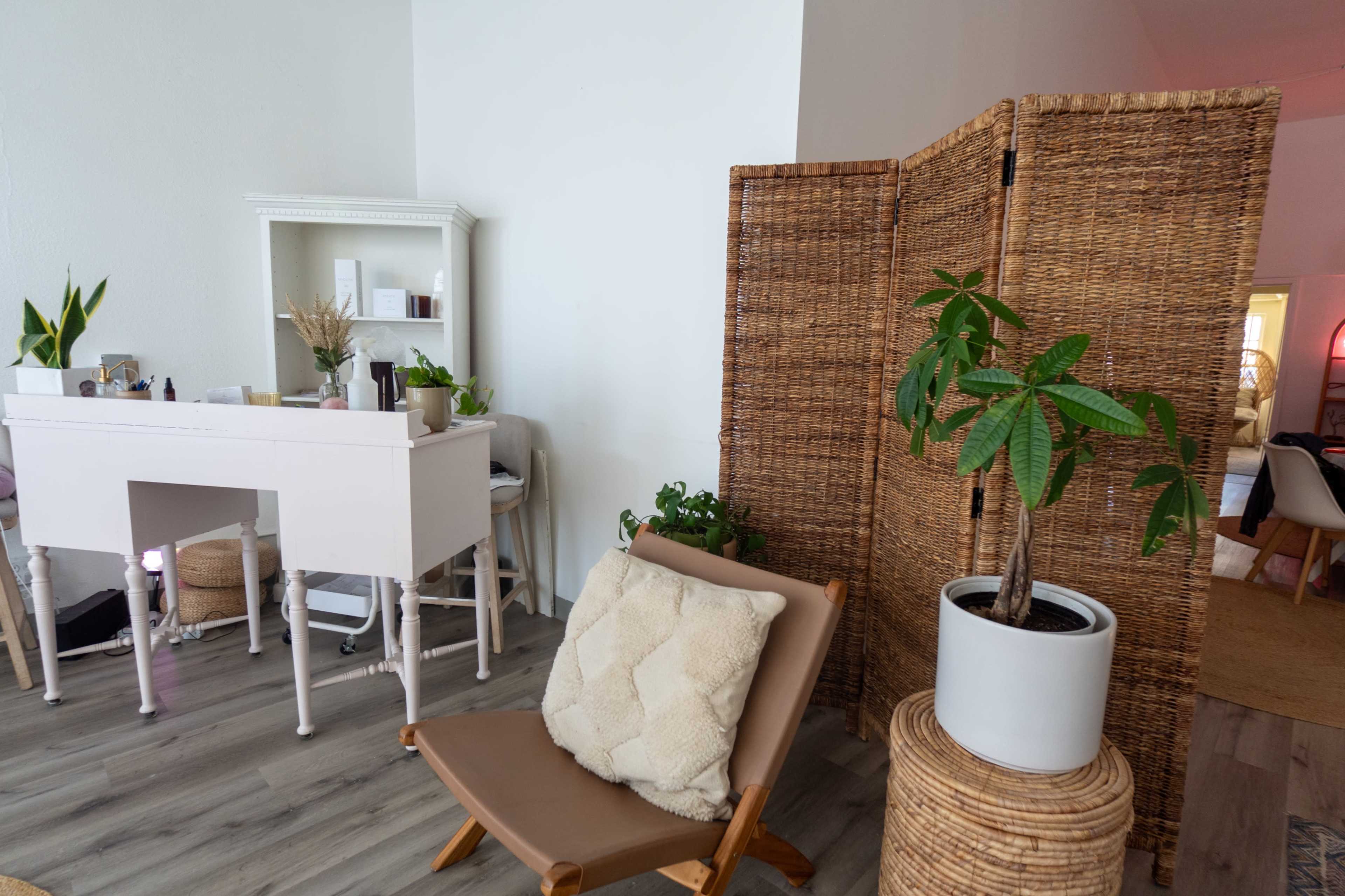 The image shows a minimalistic interior space with a white vanity, a wicker room divider, a potted plant, and a lounge chair.