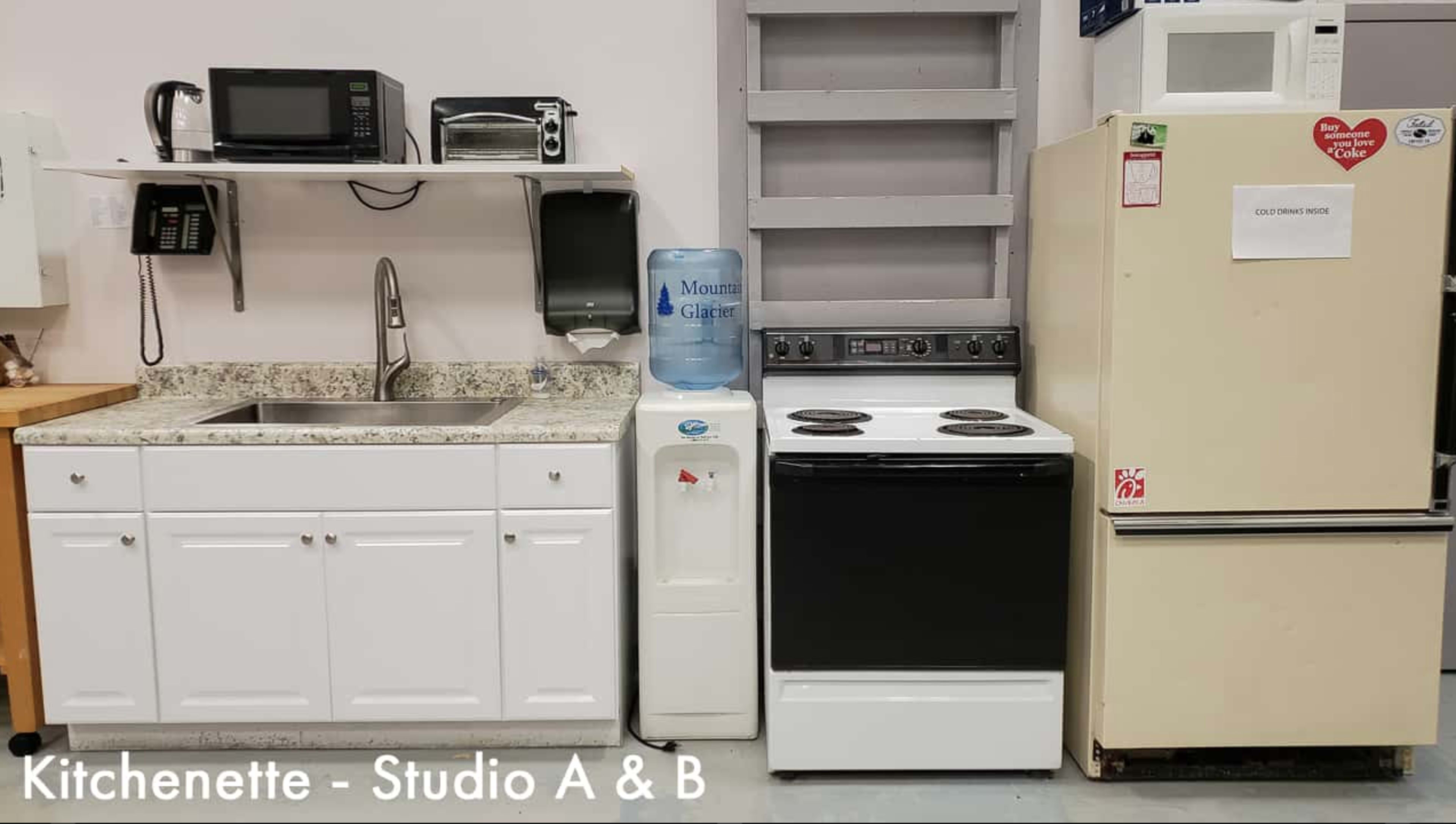 A compact kitchenette with a sink, water cooler, stove, microwave, and a refrigerator lined up against a wall.
