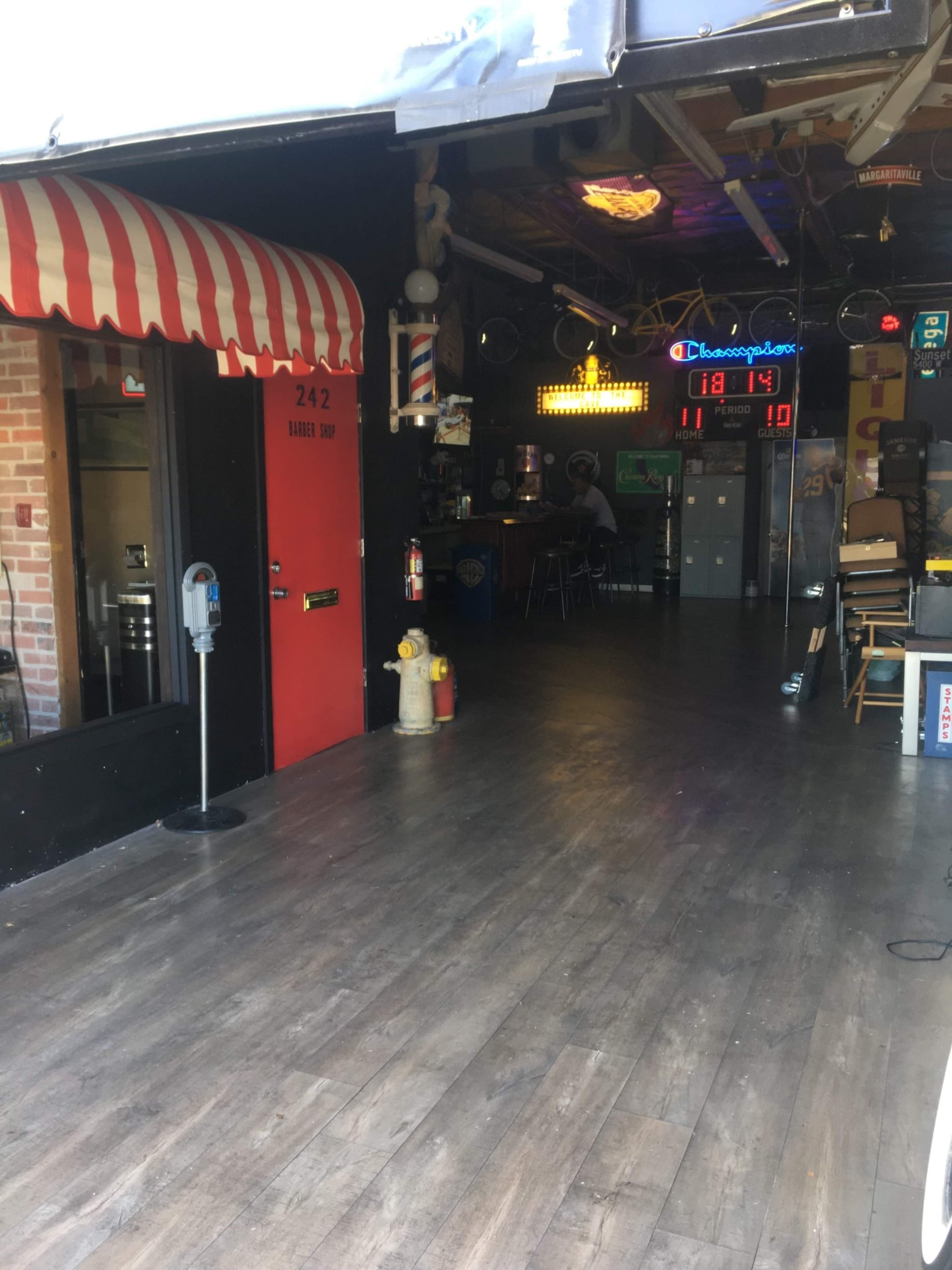 The entrance to a barbershop with a red door and a striped awning, leading into a dimly lit space with bar-style seating and various decorations.
