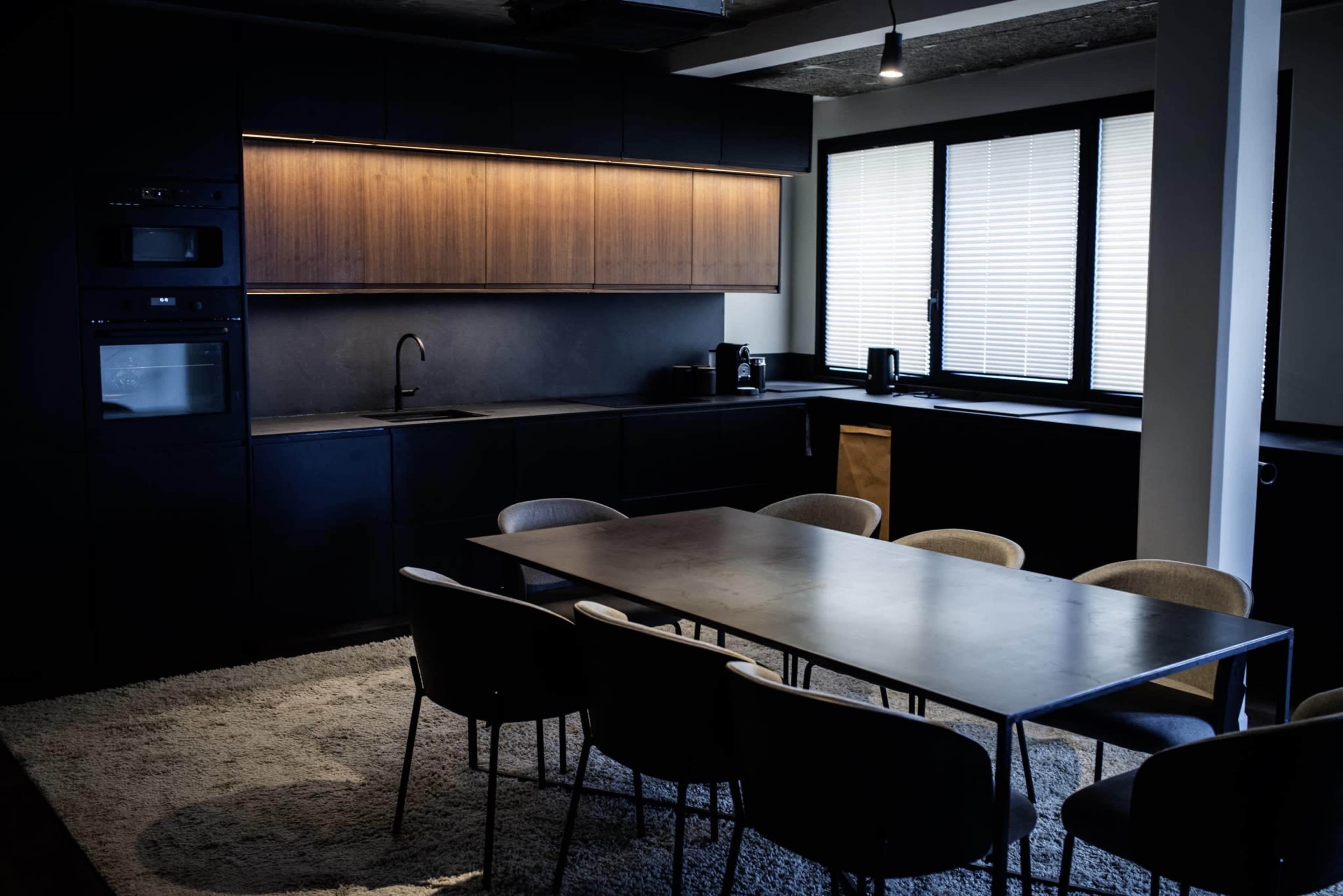 The image shows a modern kitchen with dark cabinetry, a large dining table, and a row of windows featuring horizontal blinds.