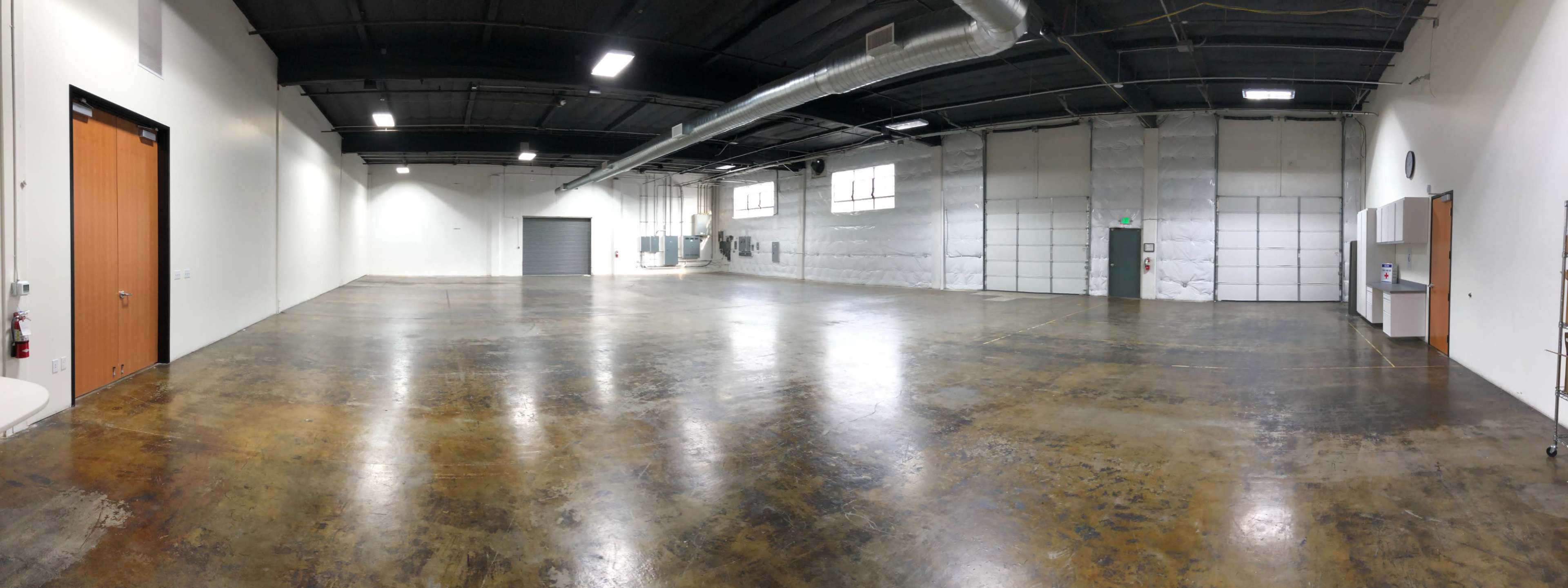 A large, empty industrial warehouse space with concrete flooring and metal overhead lighting.