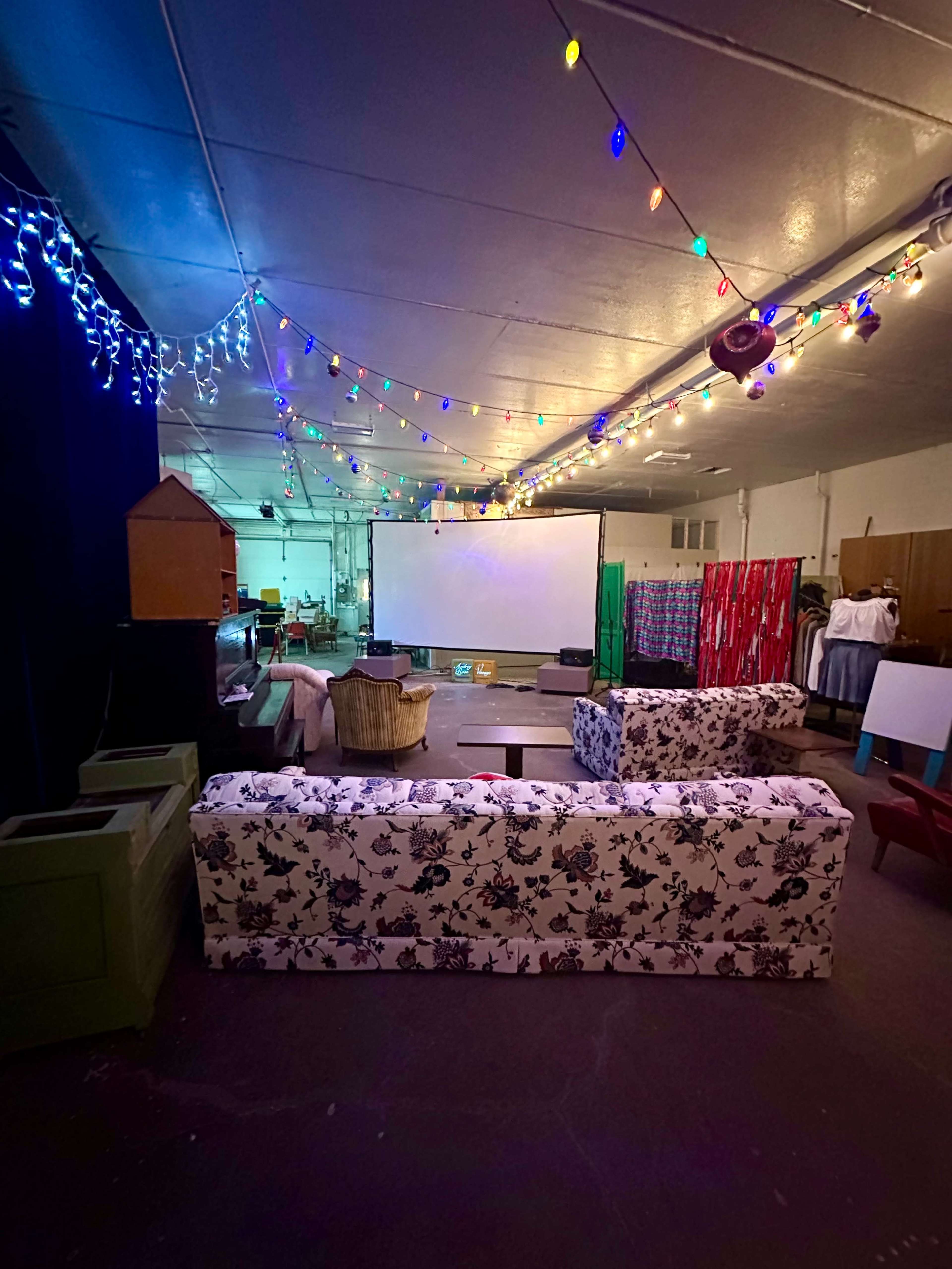 The image shows a spacious room with colorful string lights hanging from the ceiling, a large projection screen at the front, and various couches arranged in a casual seating area.