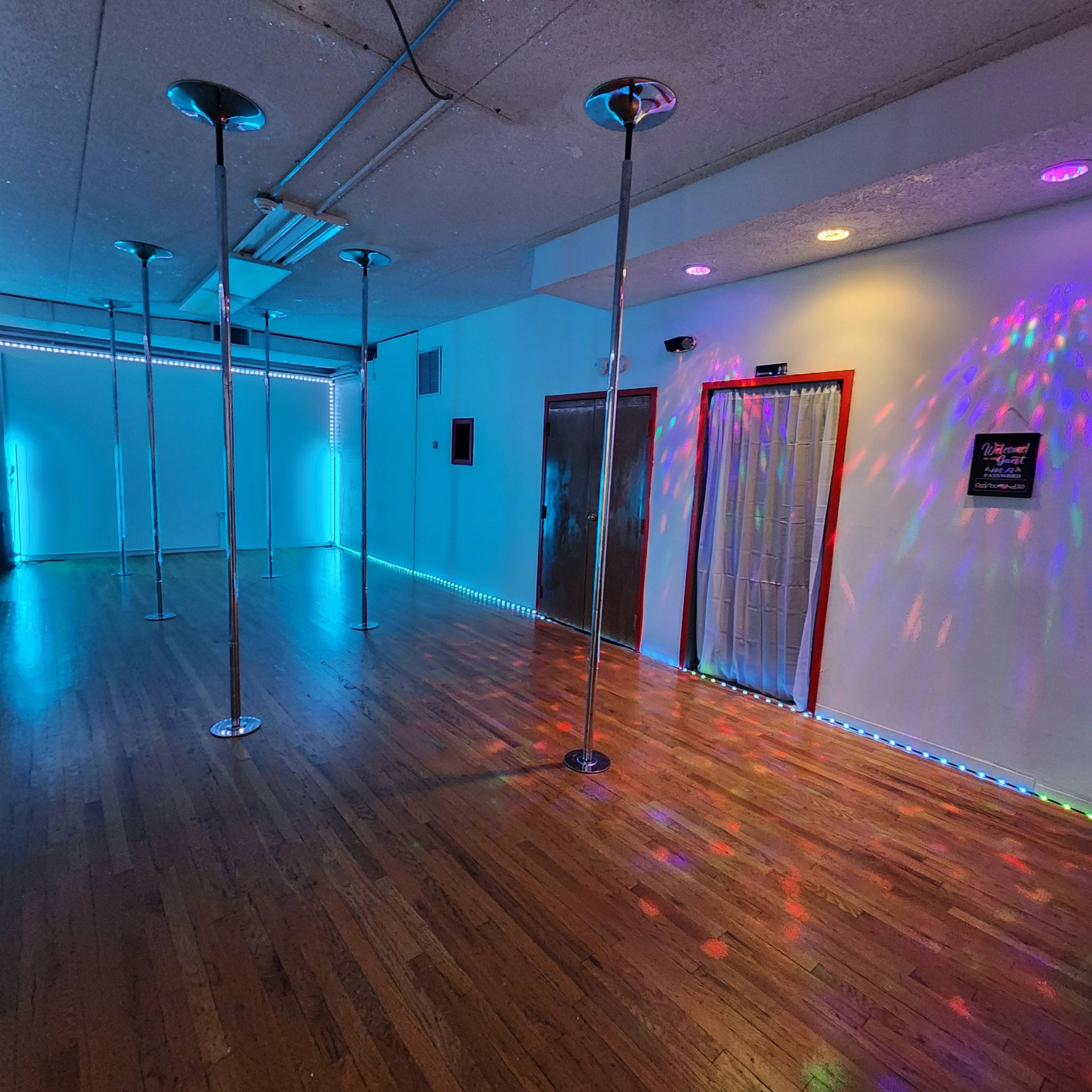 The image shows a spacious dance studio with several polished poles and colorful lighting effects on the walls and floor.