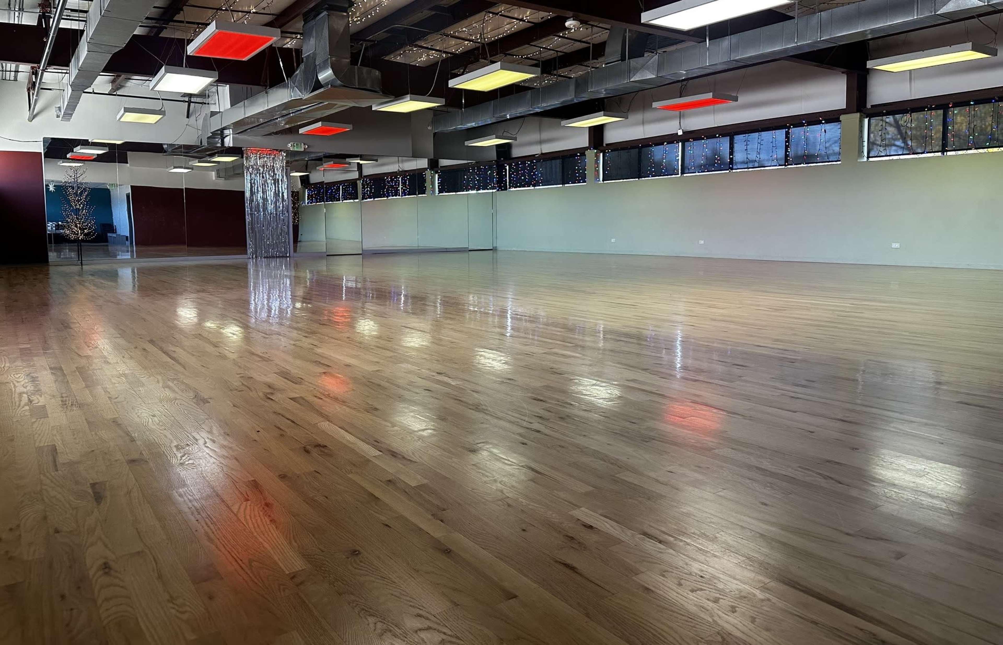 A spacious room with polished wooden floors, large windows, and suspended ceiling lights of various colors.