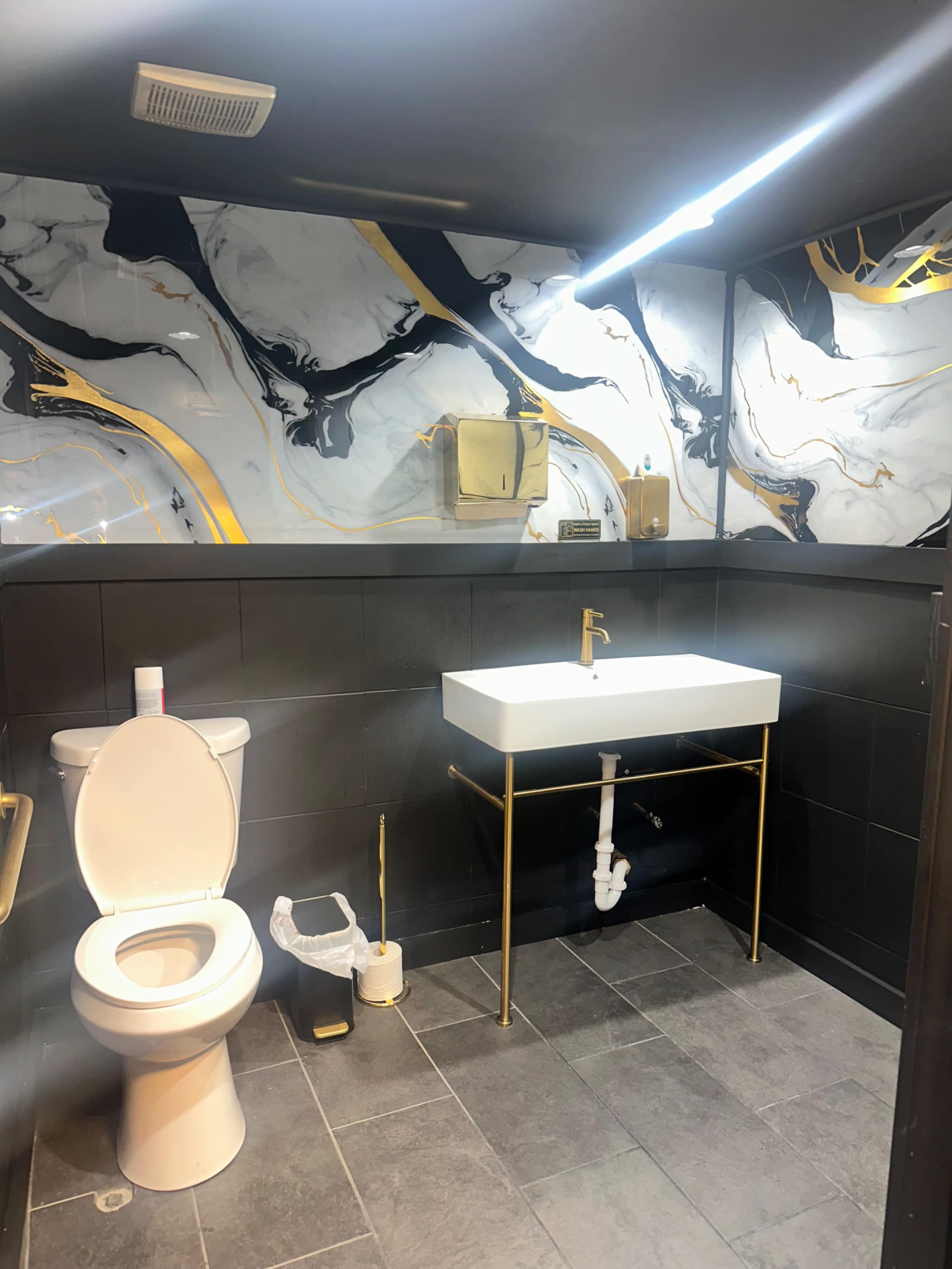 The image shows a modern bathroom featuring a gold and white marble wall design, a wall-mounted sink with a gold frame, a toilet, and a stylish towel holder.