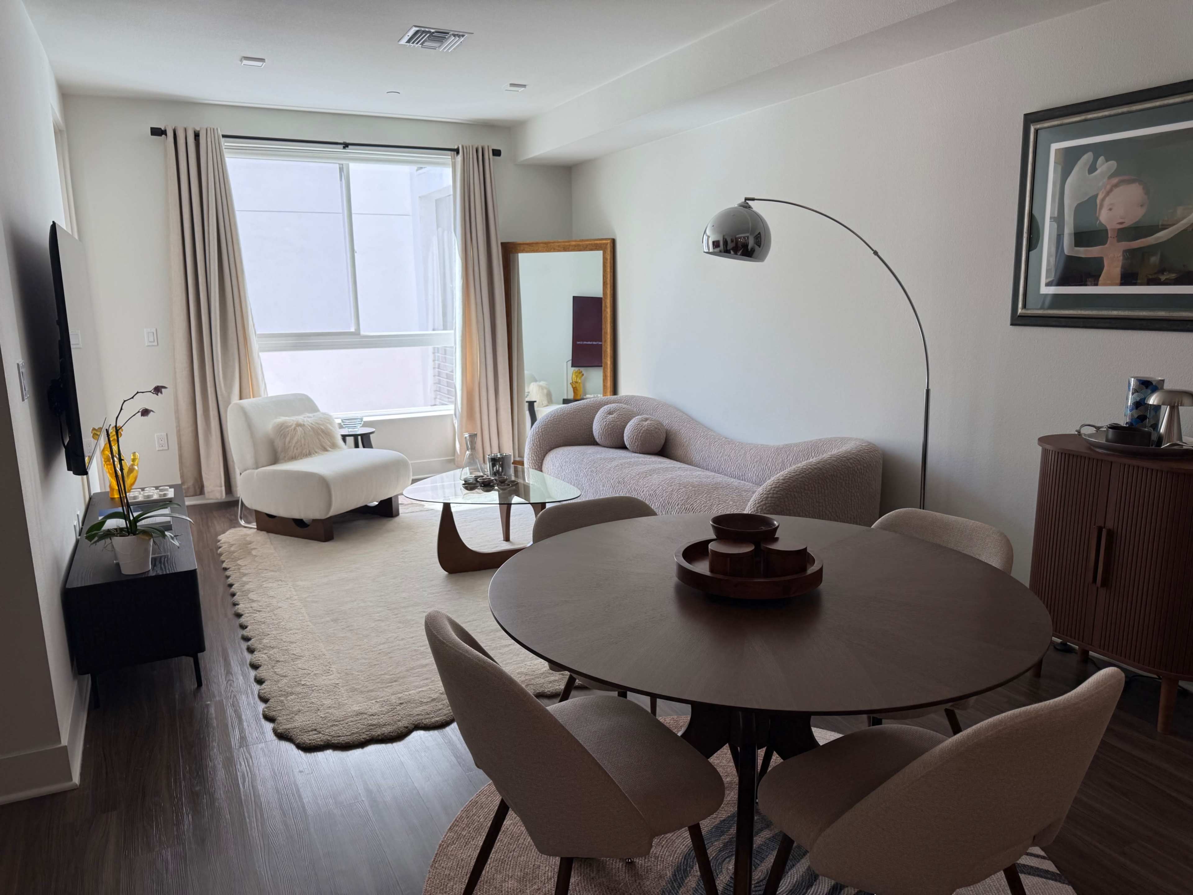 The image shows a modern, open living space featuring a round dining table, a beige couch, a large window with curtains, and decorative elements like a floor lamp and artwork.