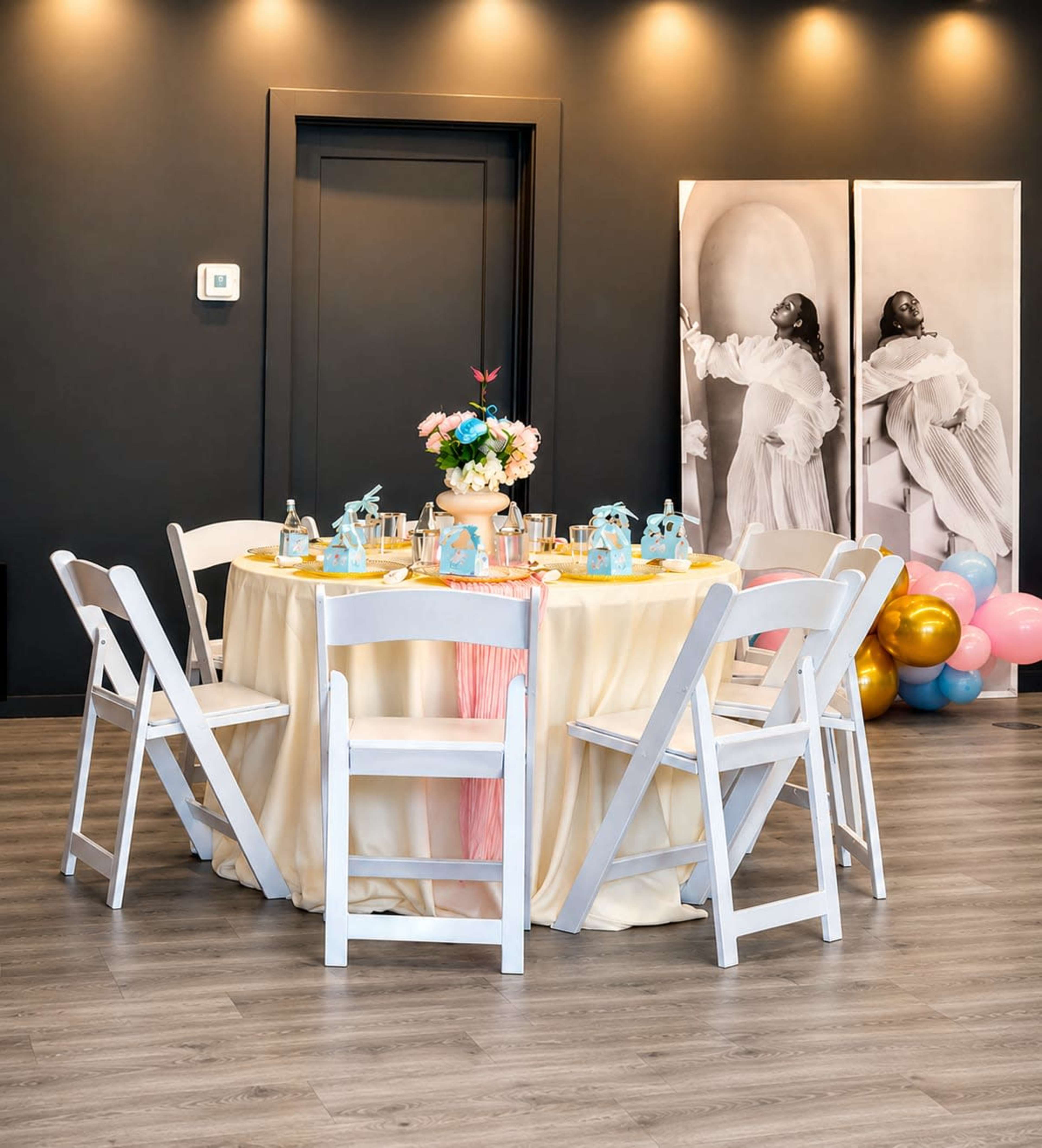 A round table draped in a light beige cloth is set with colorful party decorations and surrounded by white chairs in a stylish indoor space.