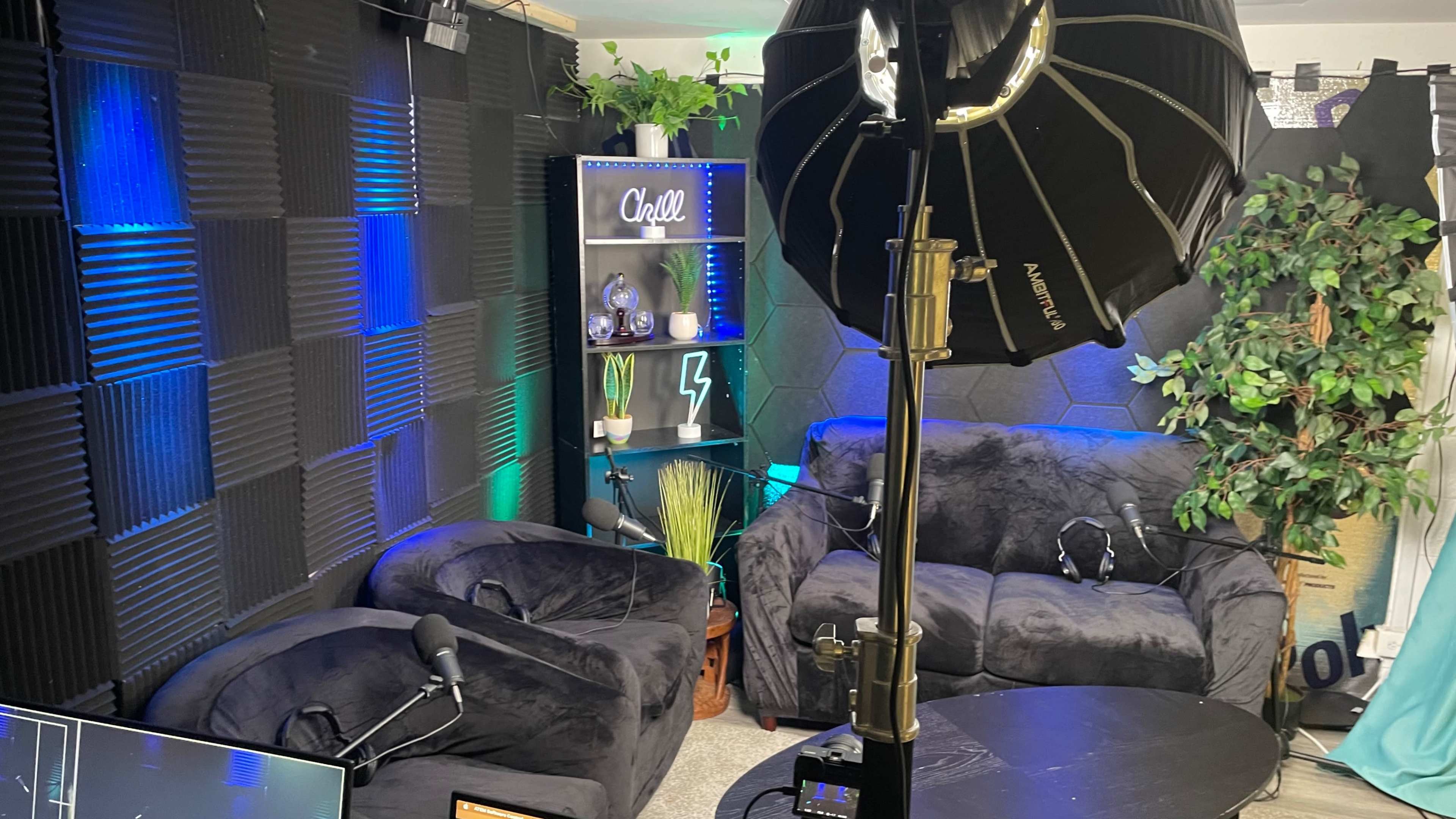 The image shows a cozy studio setup featuring two black bean bag chairs, a round table, a large softbox light, and a backdrop with acoustic panels and decorative plants.