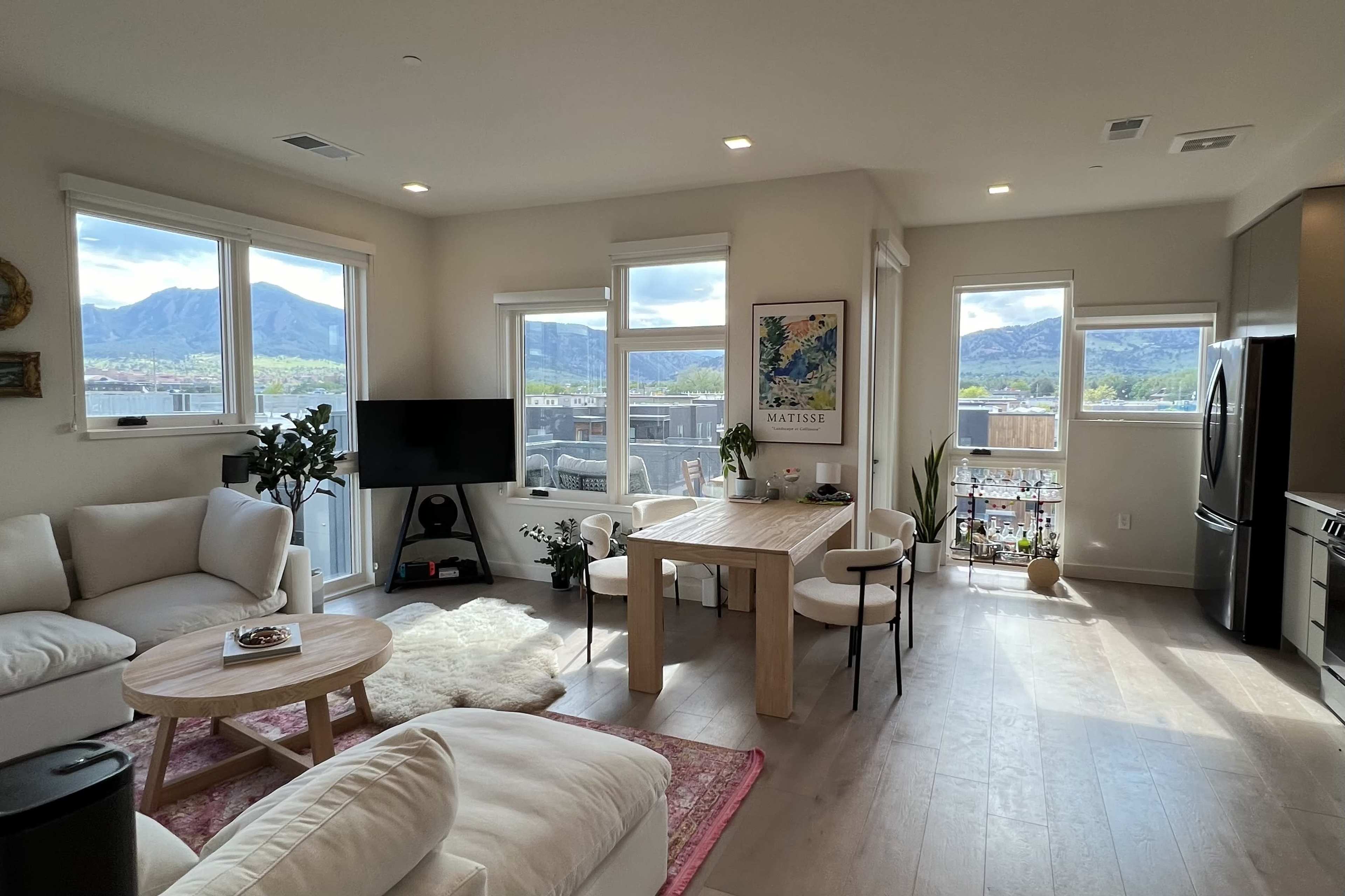 A modern living room and dining area with large windows showcasing a mountain view, featuring a white sofa, a wooden dining table, and a television.