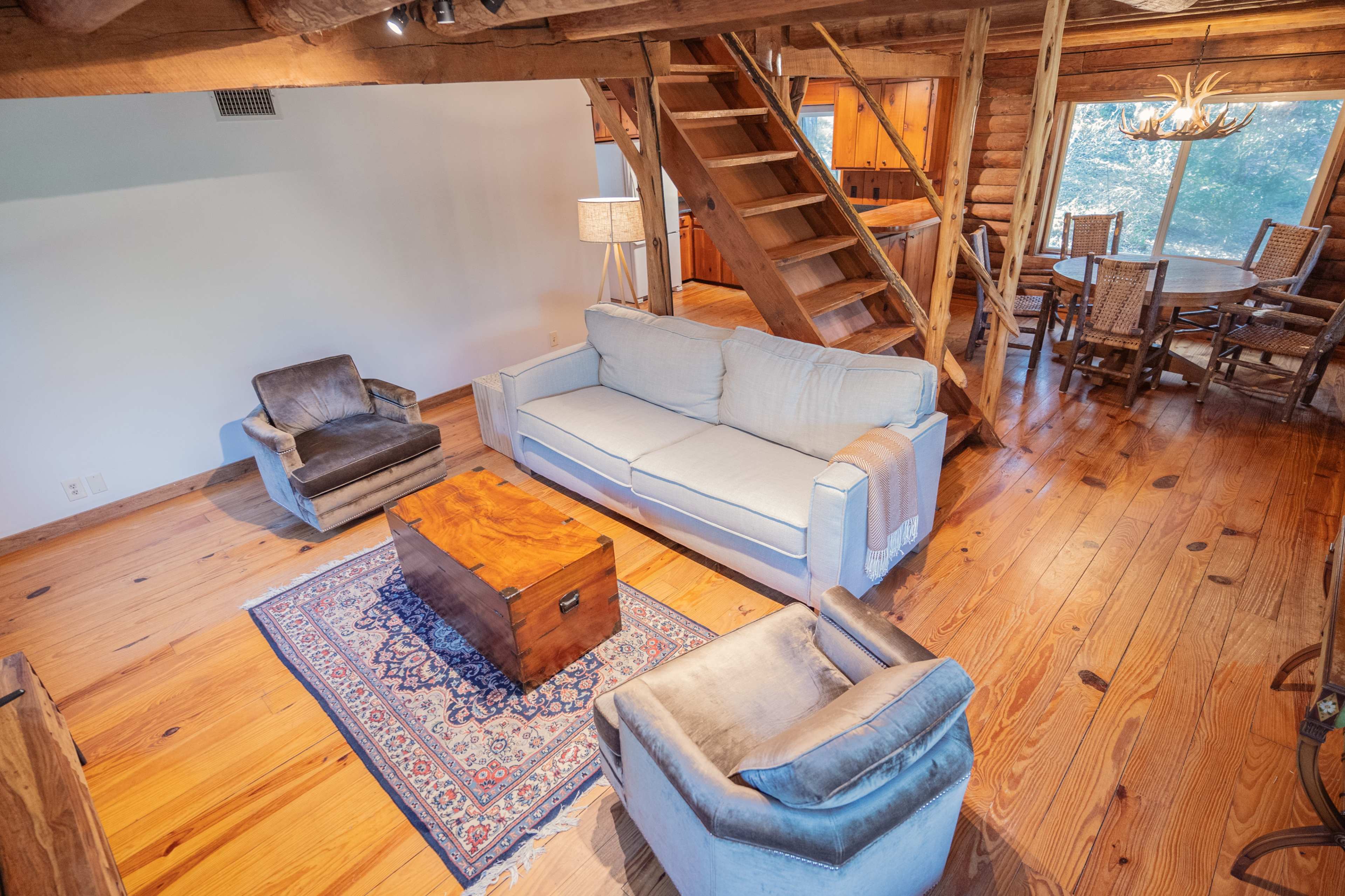 A cozy living area with a sofa, two armchairs, a wooden coffee table, and a staircase leading to an upper level, accented by wooden beams and a dining area in the background.