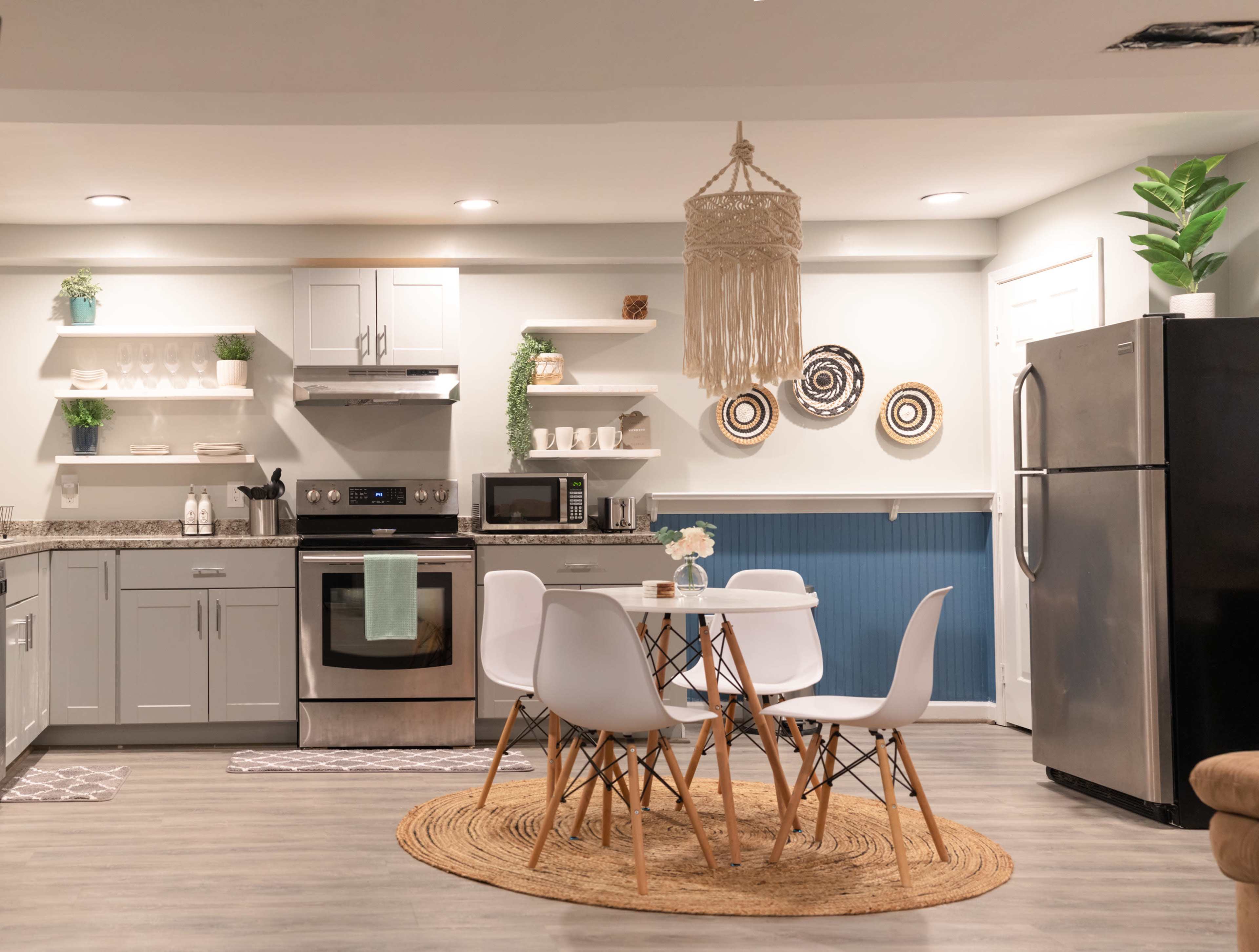 A modern kitchen features gray cabinets, stainless steel appliances, a round dining table surrounded by white chairs, and decorative elements like wall shelves and plants.