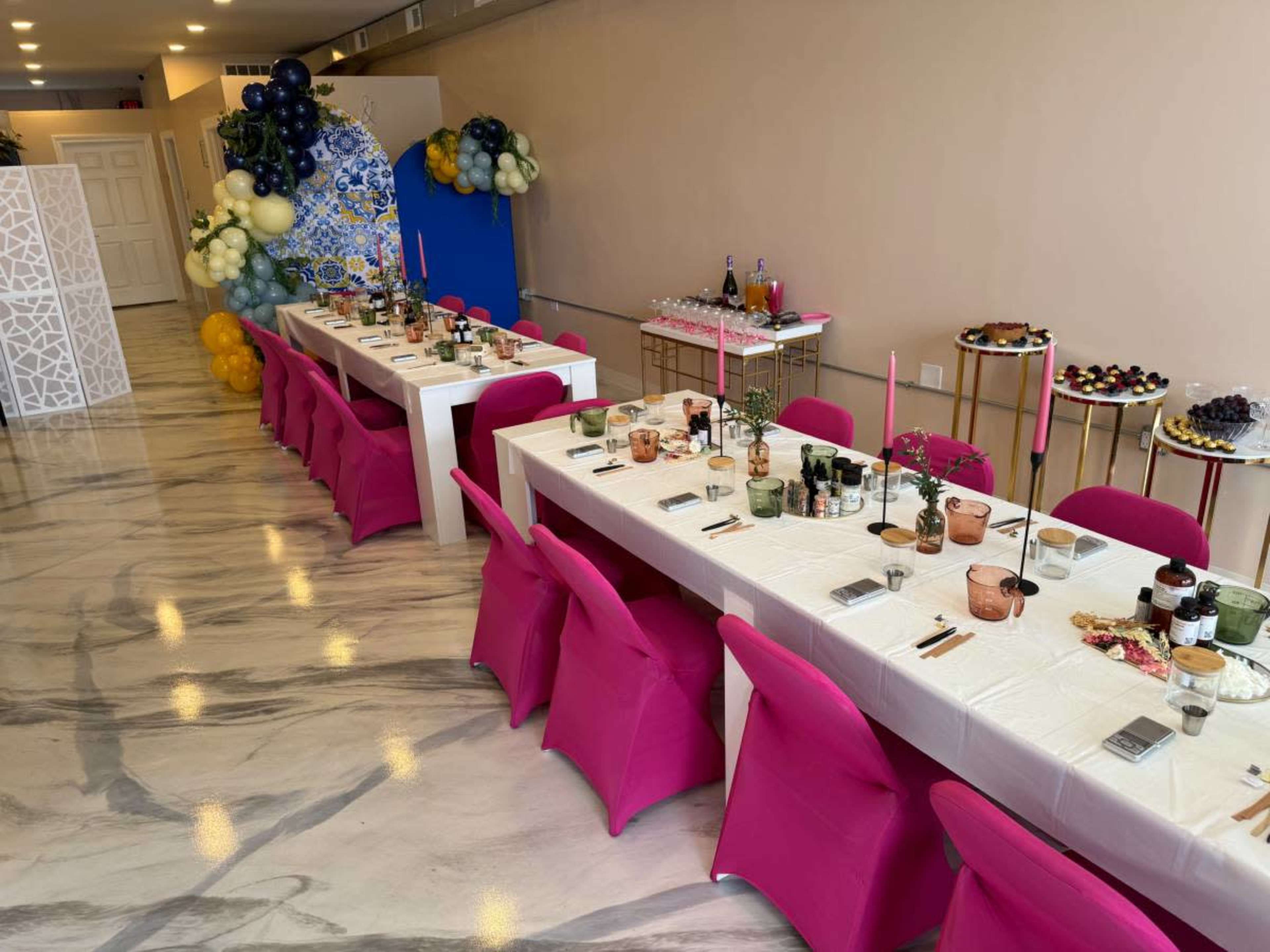 A decorated event space with two long tables set for dining, featuring pink chairs, colorful candles, and an array of food displays nearby.