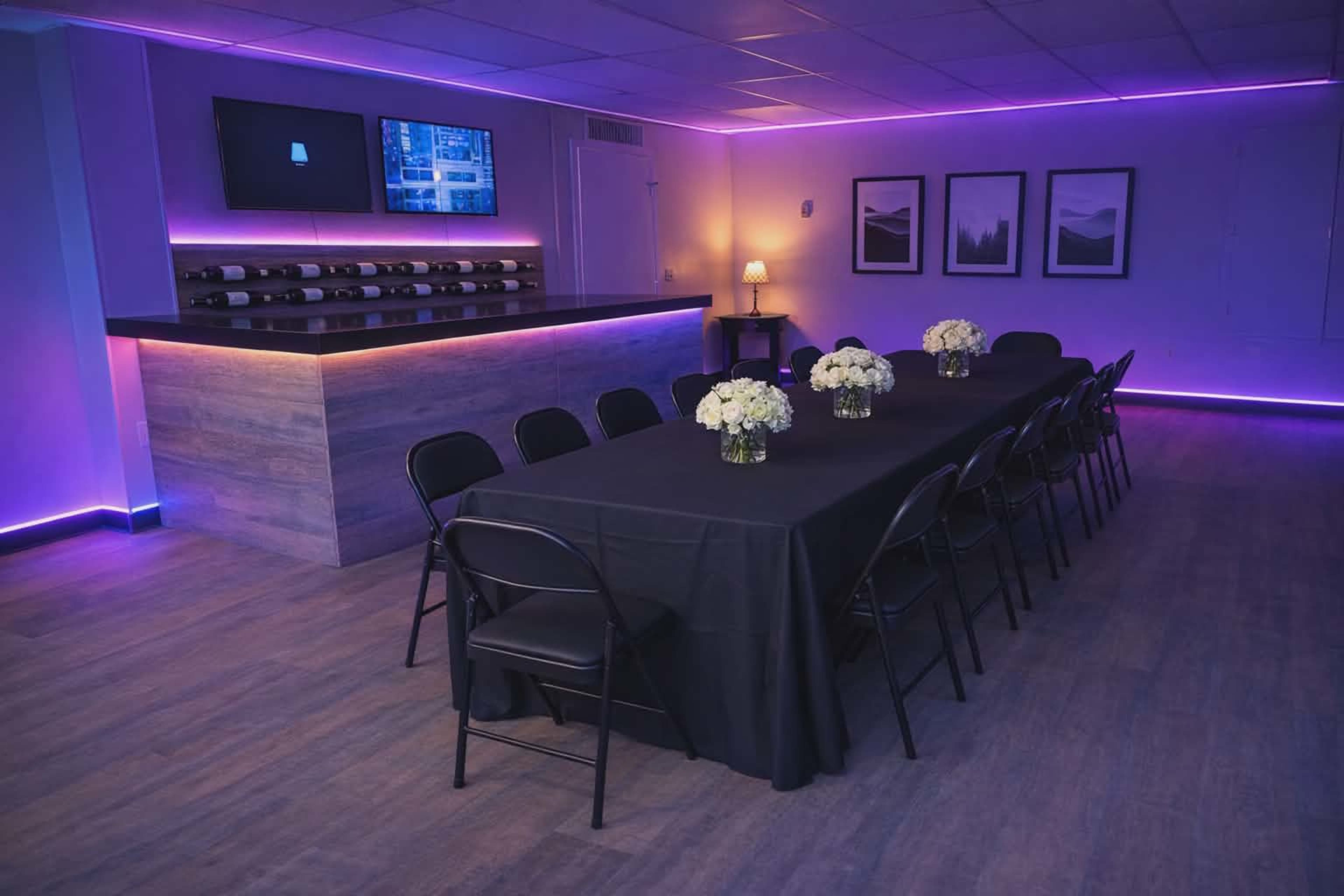A long black table is set for dining in a room with purple LED lighting, featuring a wine rack and two framed pictures on the wall.