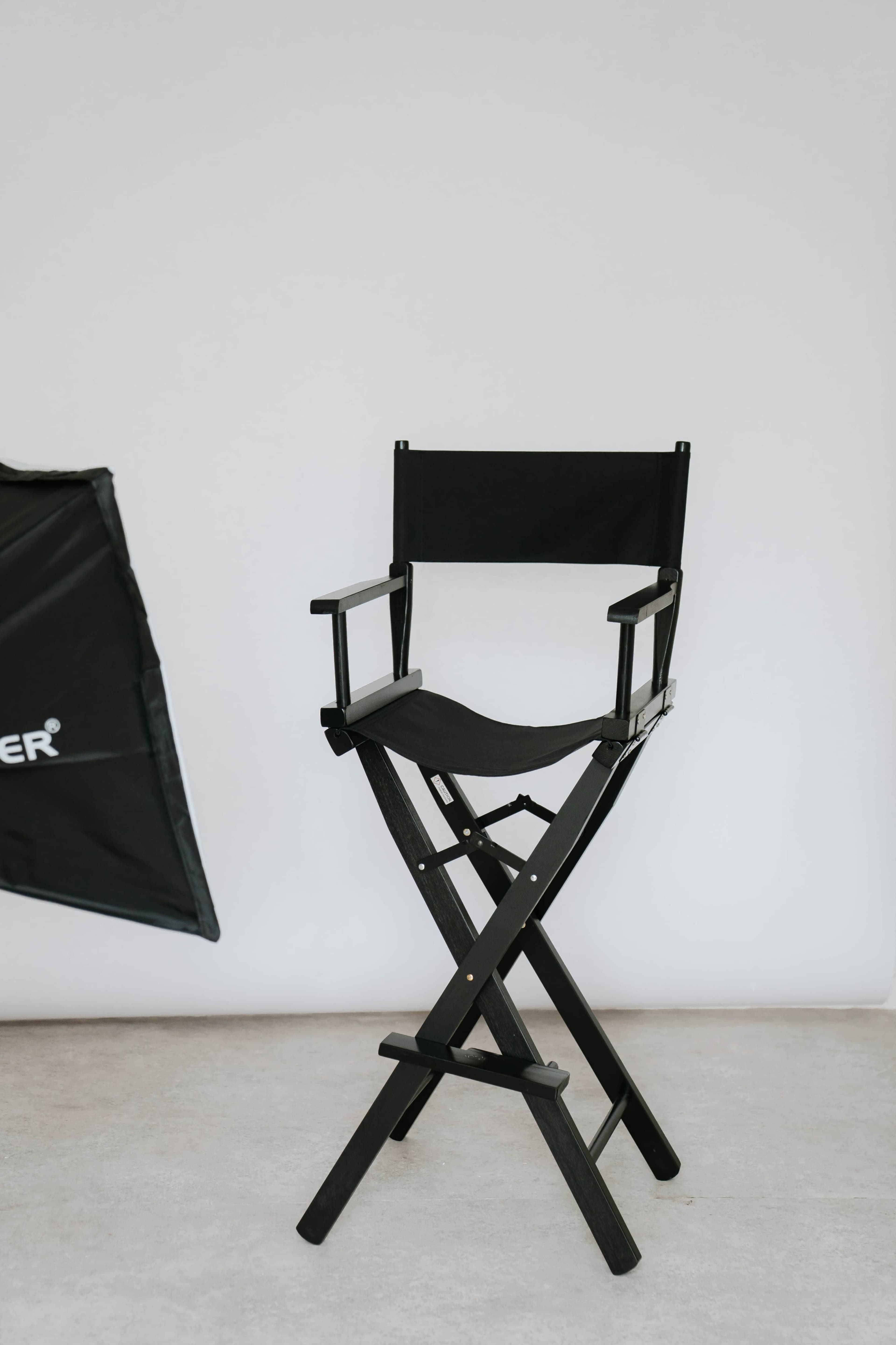 A black director's chair stands against a plain white wall, with a photography umbrella visible to the side.