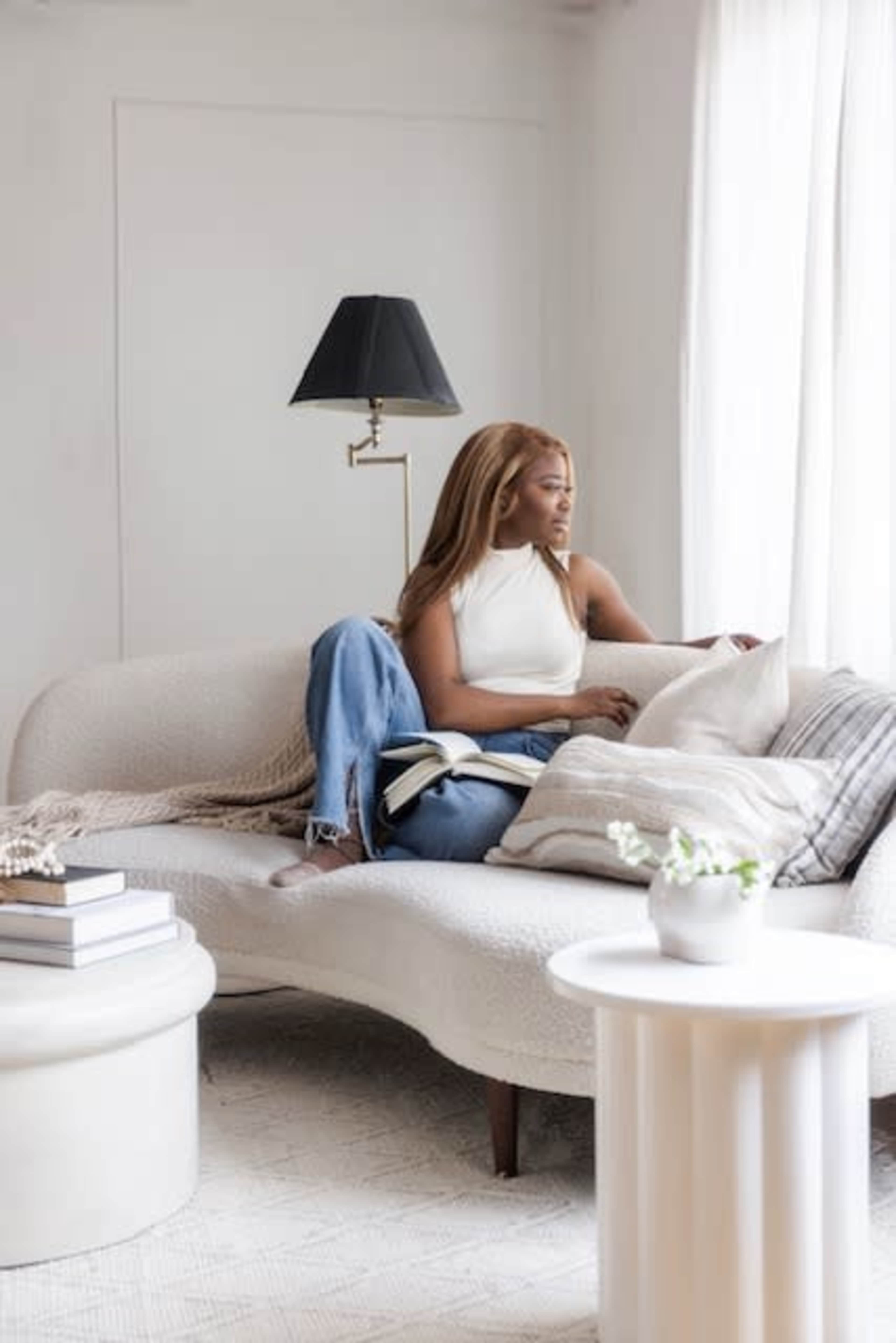 A woman sits on a curved white sofa in a bright room, looking out the window while holding a book.