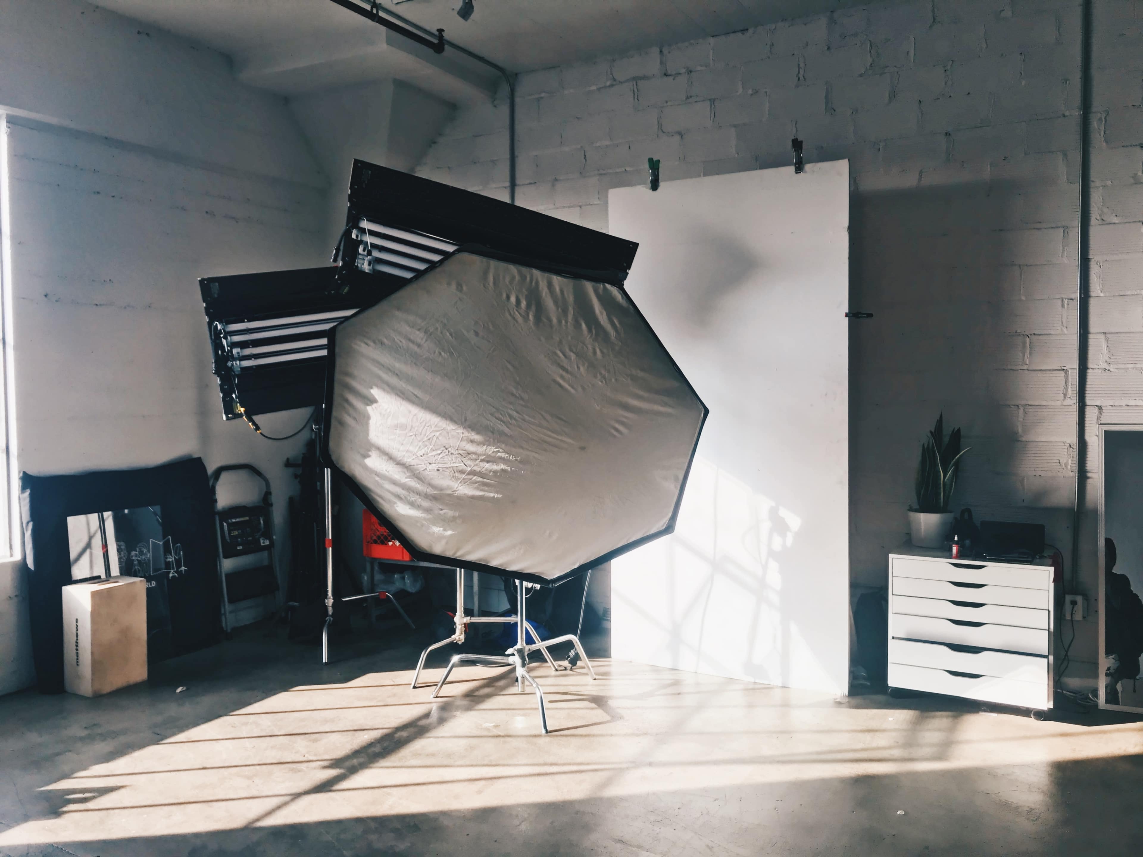 A photography studio features an octagonal softbox, a white backdrop, and a low dresser, illuminated by sunlight streaming through the windows.