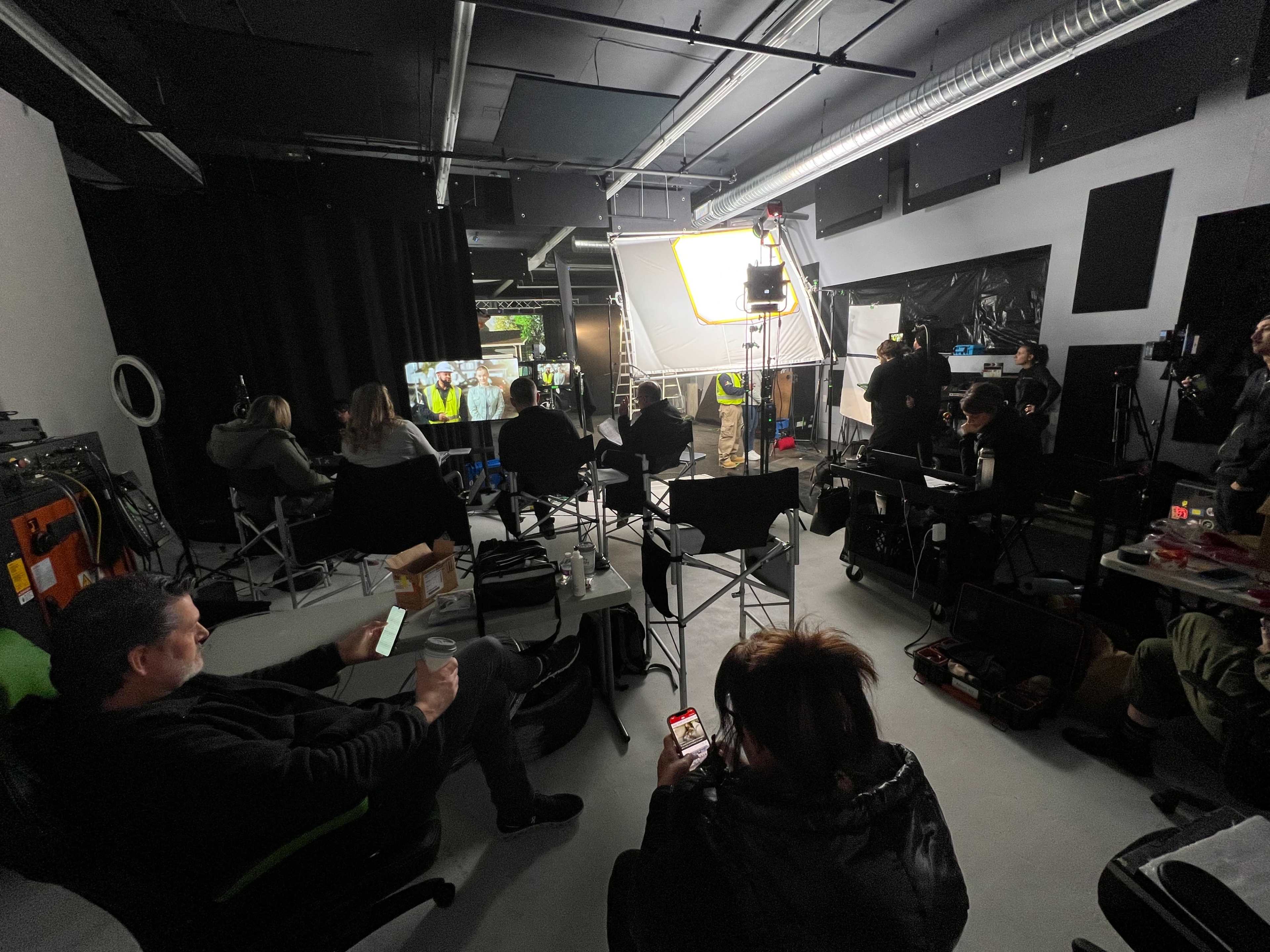 A film crew is gathered in a studio while a scene is being filmed, with various equipment and monitors visible.