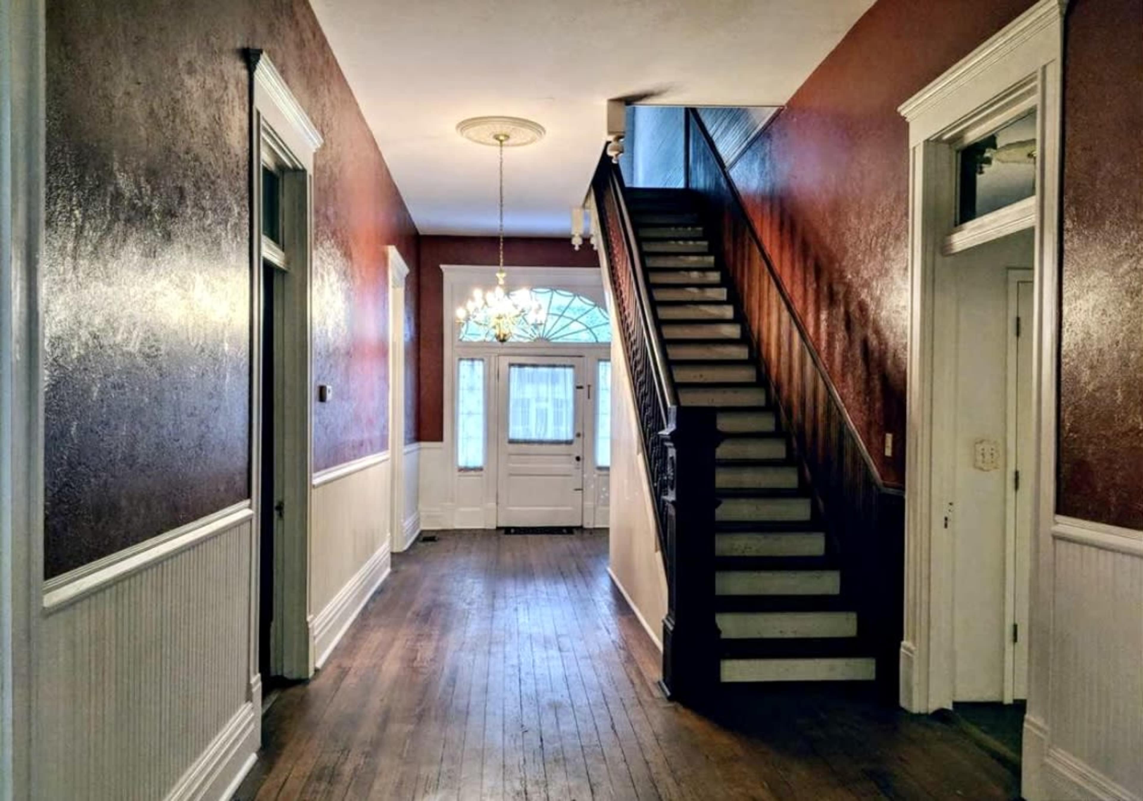 The image shows a hallway with wooden flooring, white wainscoting, a staircase on one side, and a chandelier hanging from the ceiling.