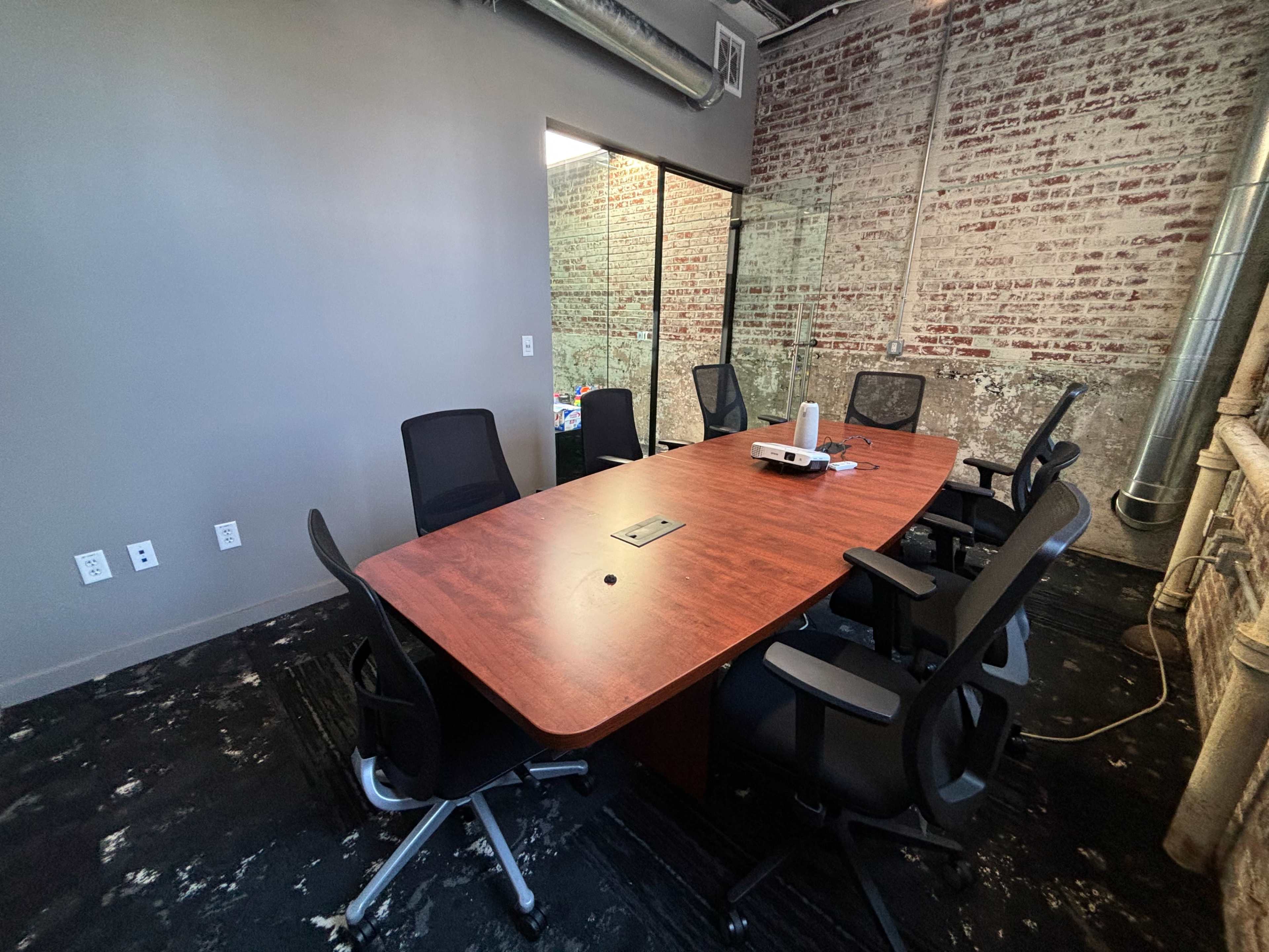 A rectangular wooden conference table surrounded by six black office chairs is set up in a room with exposed brick walls and a glass partition.